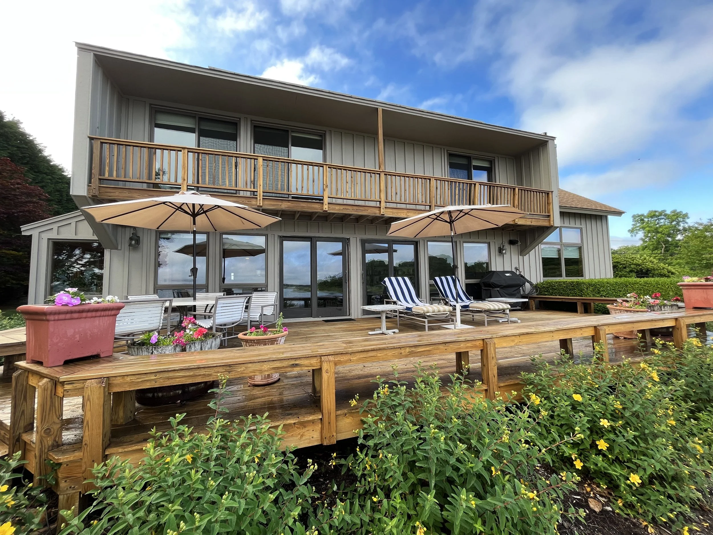 A two-story house with a wooden deck, outdoor seating, umbrellas, potted plants, and a grill, overlooking a garden and a body of water under a blue sky with clouds.