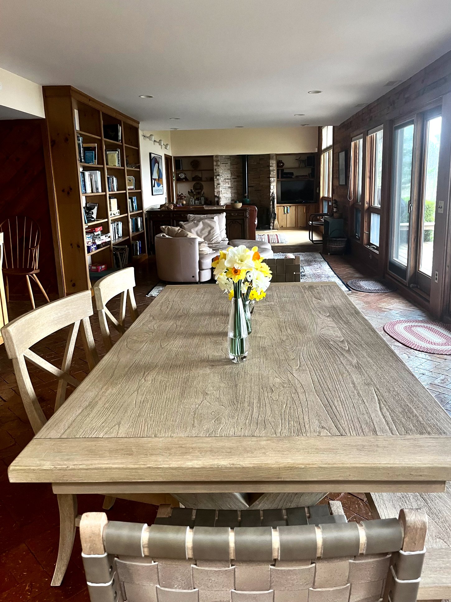 A rectangular wooden dining table with a glass vase holding yellow and white flowers, in a spacious living room with large windows, a white sofa, a bookshelf, and a fireplace.