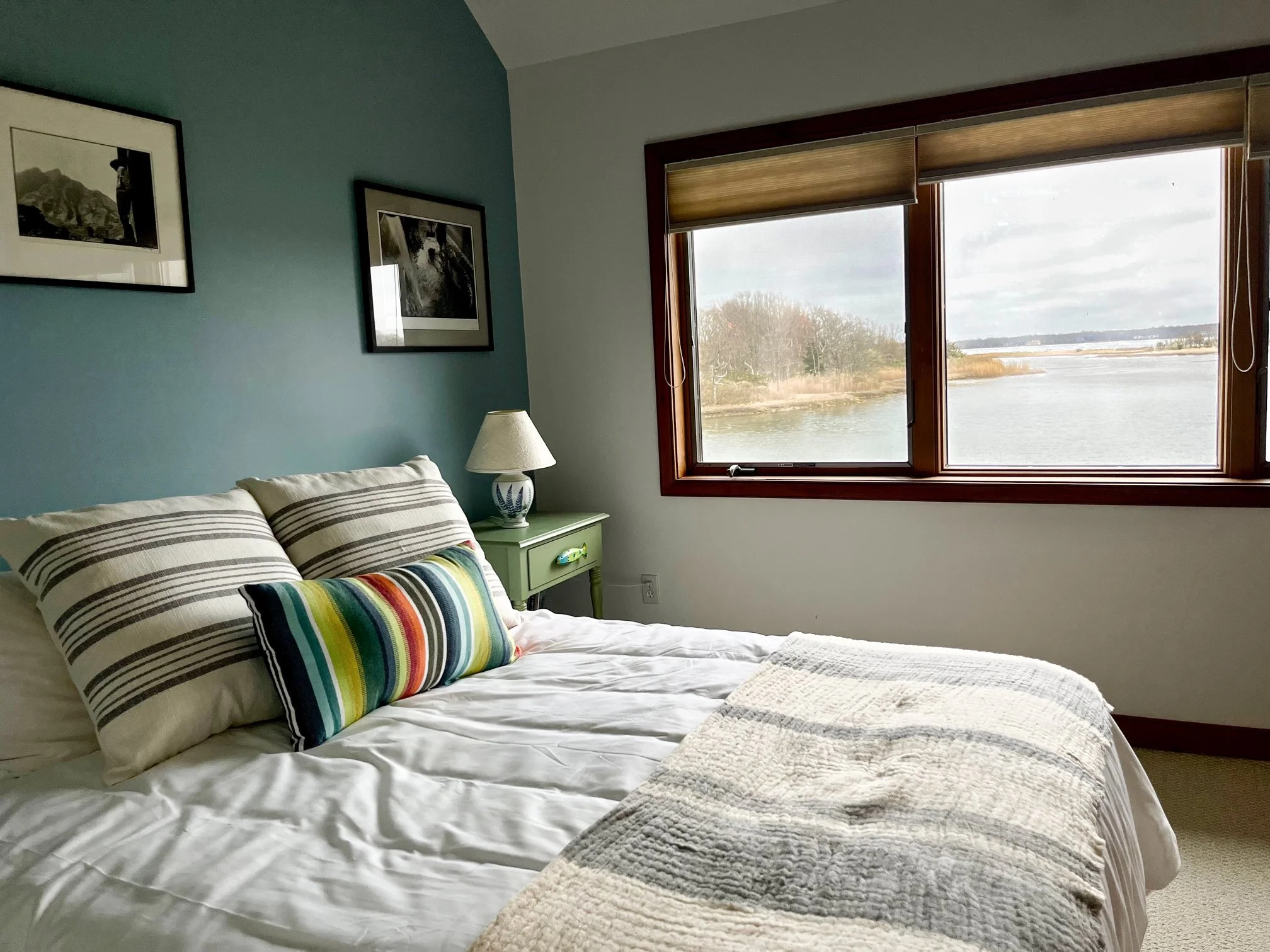 A cozy bedroom with a blue accent wall, framed black and white photographs, a window overlooking a body of water, a bedside table with a lamp, and a bed with striped pillows and a textured throw blanket.