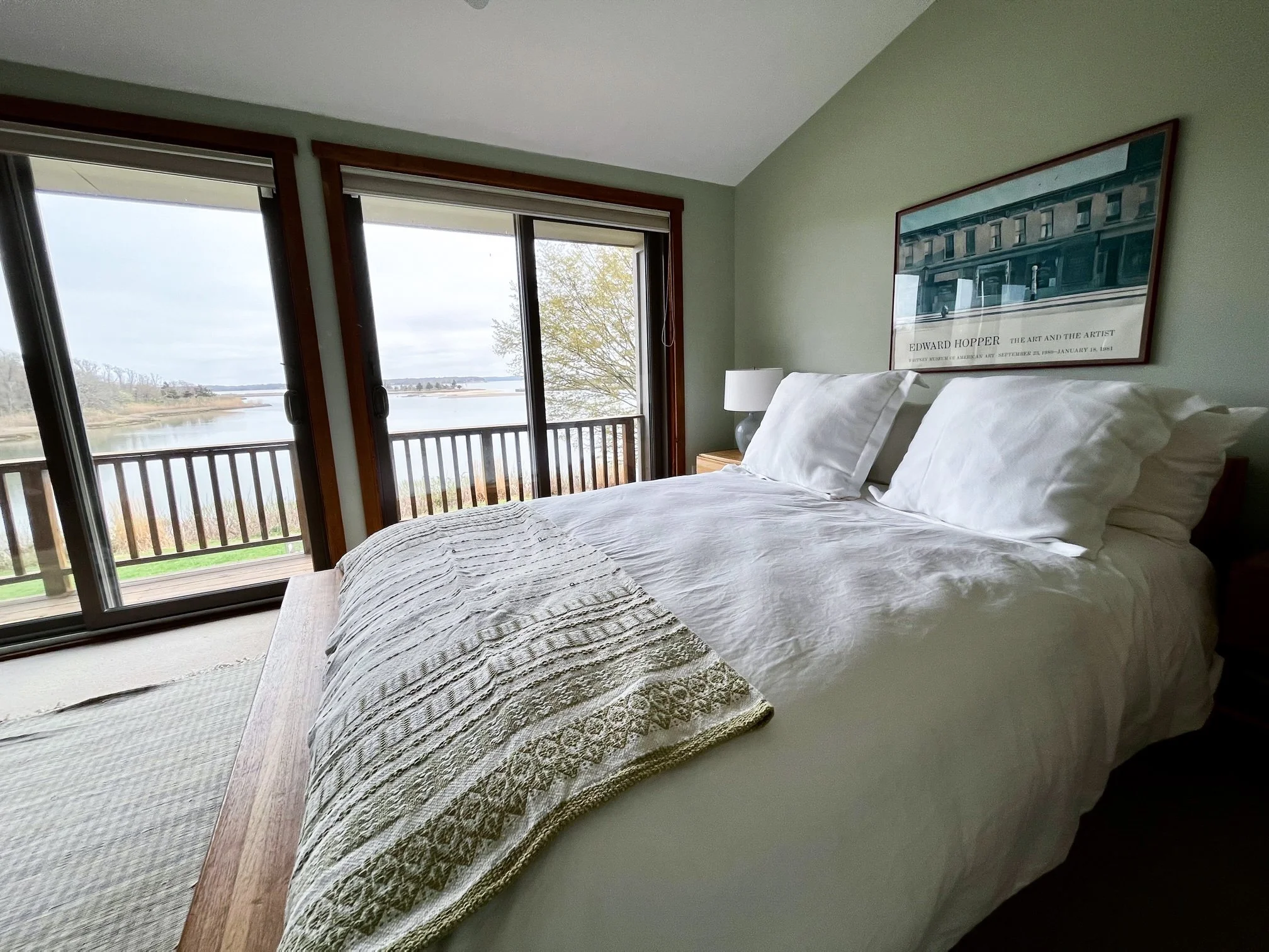 A bedroom with a bed near sliding glass doors that open to a balcony with a view of a lake or river. The bed has white linens and pillows, a patterned throw blanket at the foot, and a framed art print above.