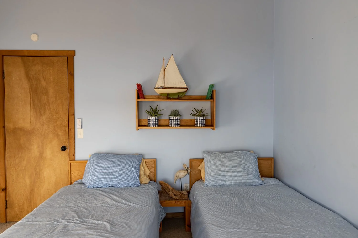 A bedroom with two beds, each with light gray bedding and pillows, a wooden nightstand with a bird sculpture, and a wooden shelf with plants and a model sailboat on the pale blue wall.