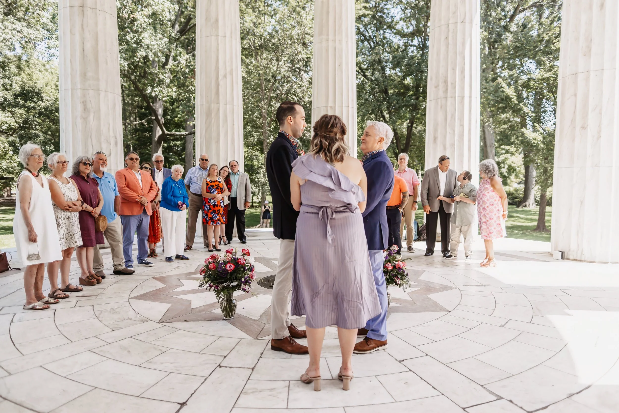 A wedding ceremony with guests gathered around a couple exchanging vows in an outdoor setting with white pillars. The guests are dressed in semi-formal attire, and there are floral arrangements on the ground.