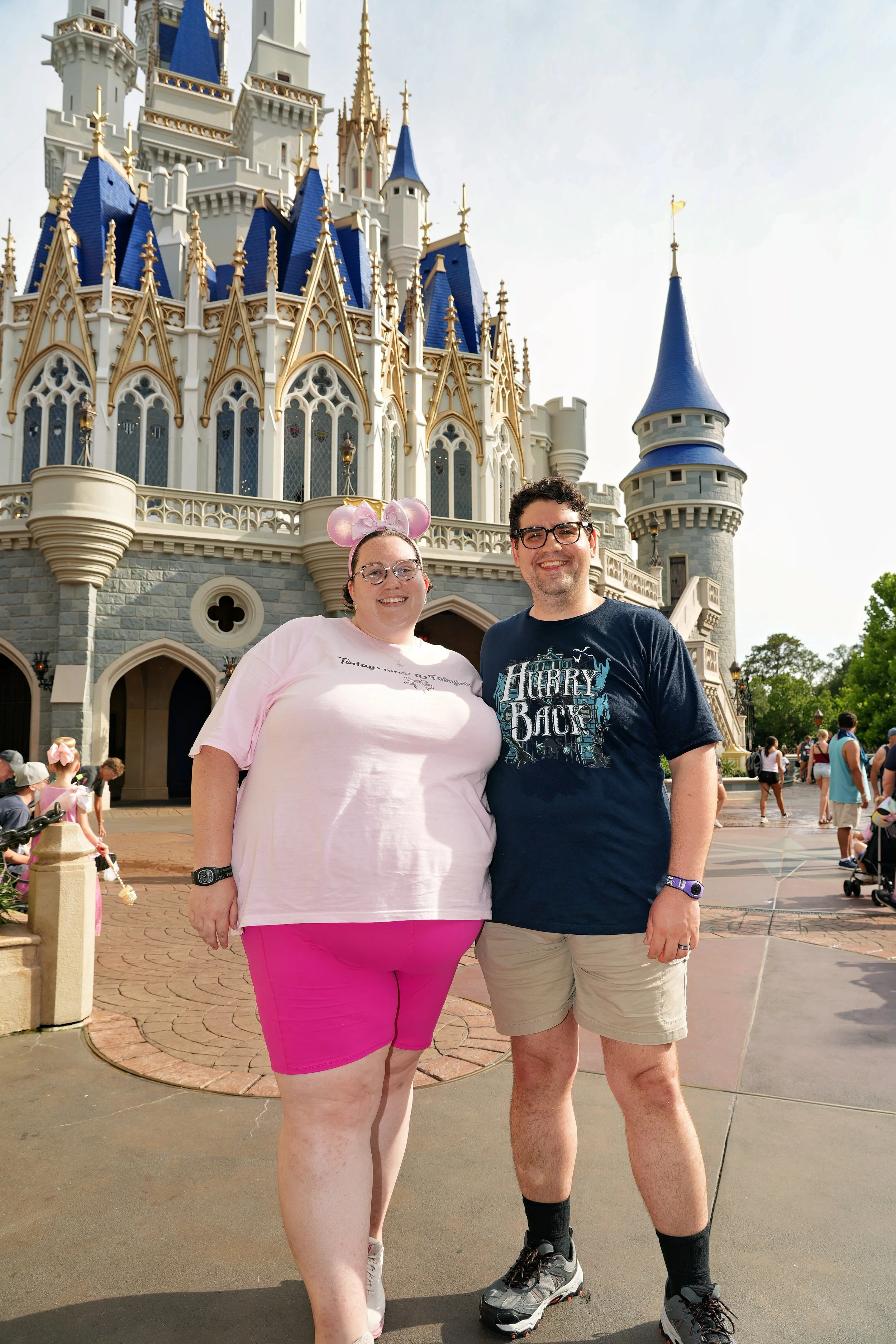 A smiling woman wearing pink shorts and a light pink t-shirt with a cartoon mouse headband, and a smiling man wearing beige shorts, a navy t-shirt, and hiking shoes,