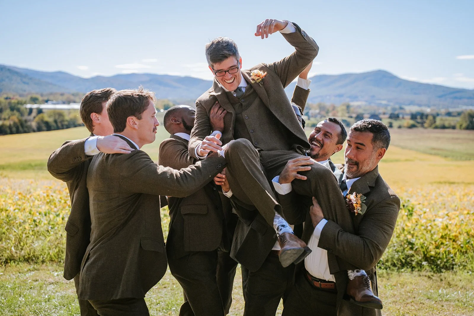 Group of men in suits lifting one man in a suit and glasses in the air outdoors, with mountains in the background, celebrating.