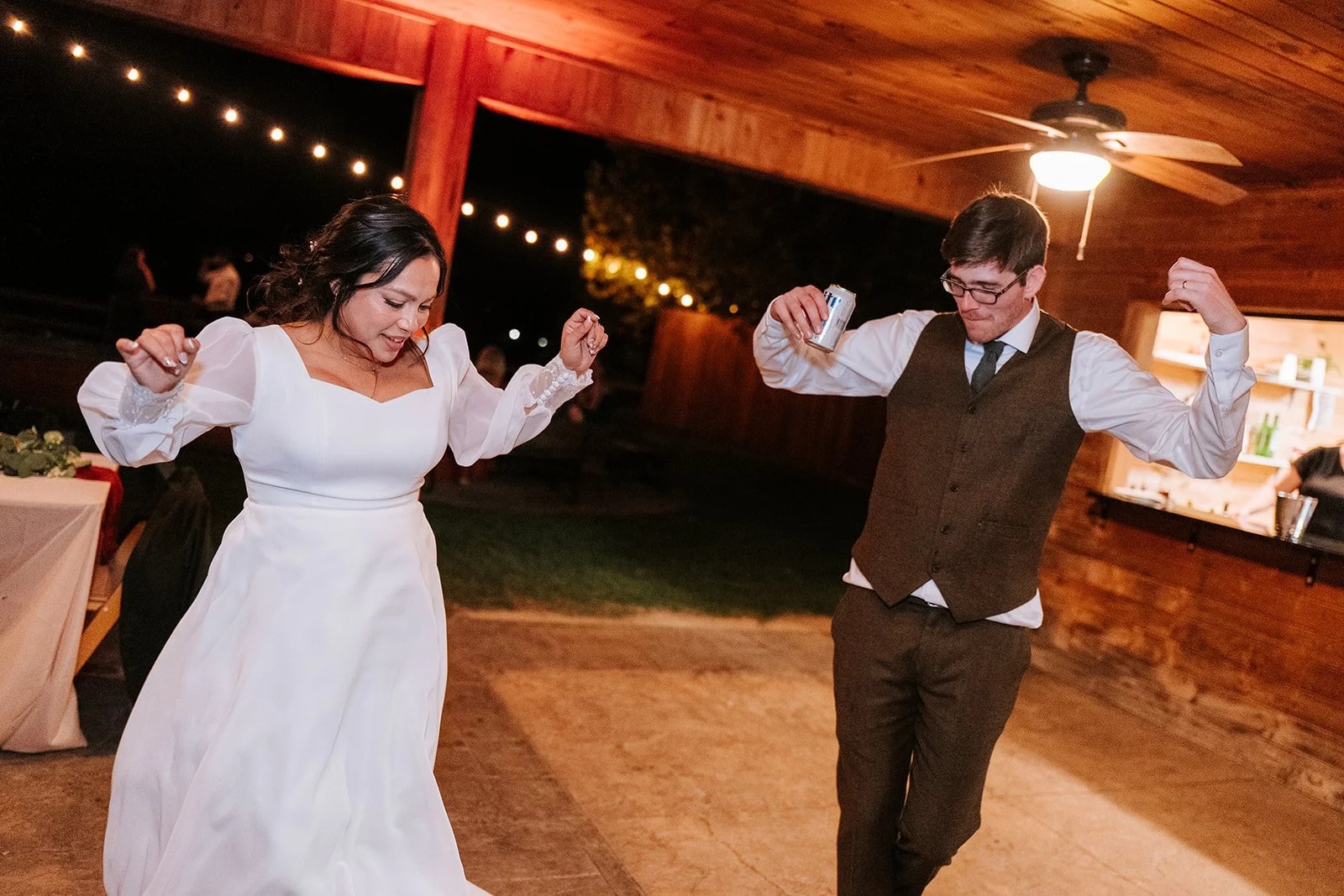 A bride and groom dancing at their wedding reception in a rustic venue with wooden walls and ceiling, decorated with string lights, at night.