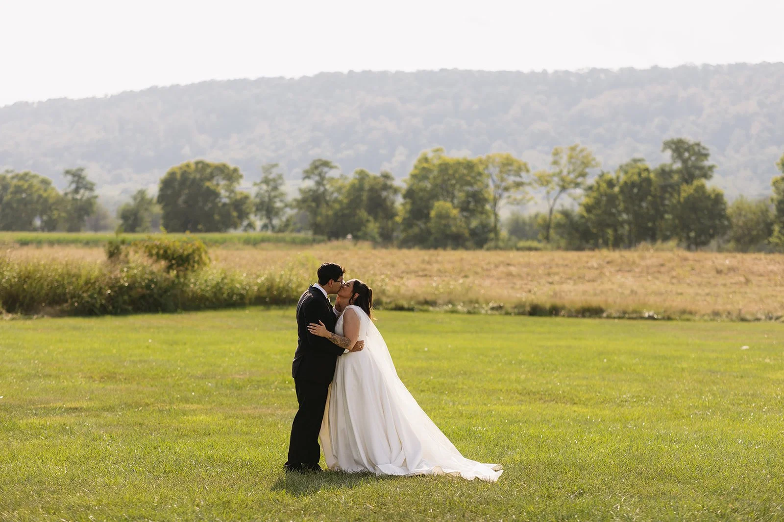 A bride and groom sharing a kiss in an open grassy field with trees and mountains in the background.