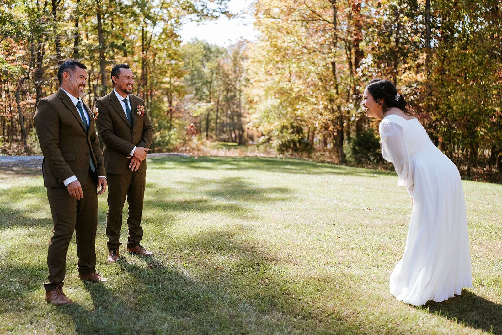 A bride in a white wedding dress leans forward smiling at two men in suits who are smiling back at her in an outdoor setting with trees and grass.