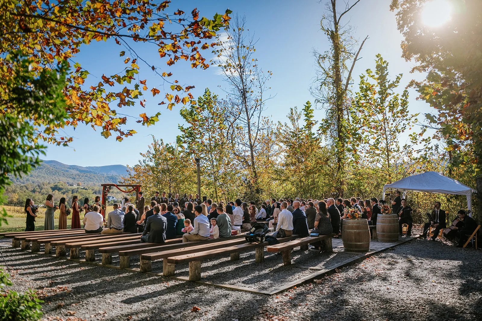 An outdoor wedding ceremony with guests seated on wooden benches, a wedding arch decorated with flowers, and a small band playing music on the right, set amidst trees with fall foliage and distant mountains under a clear blue sky.