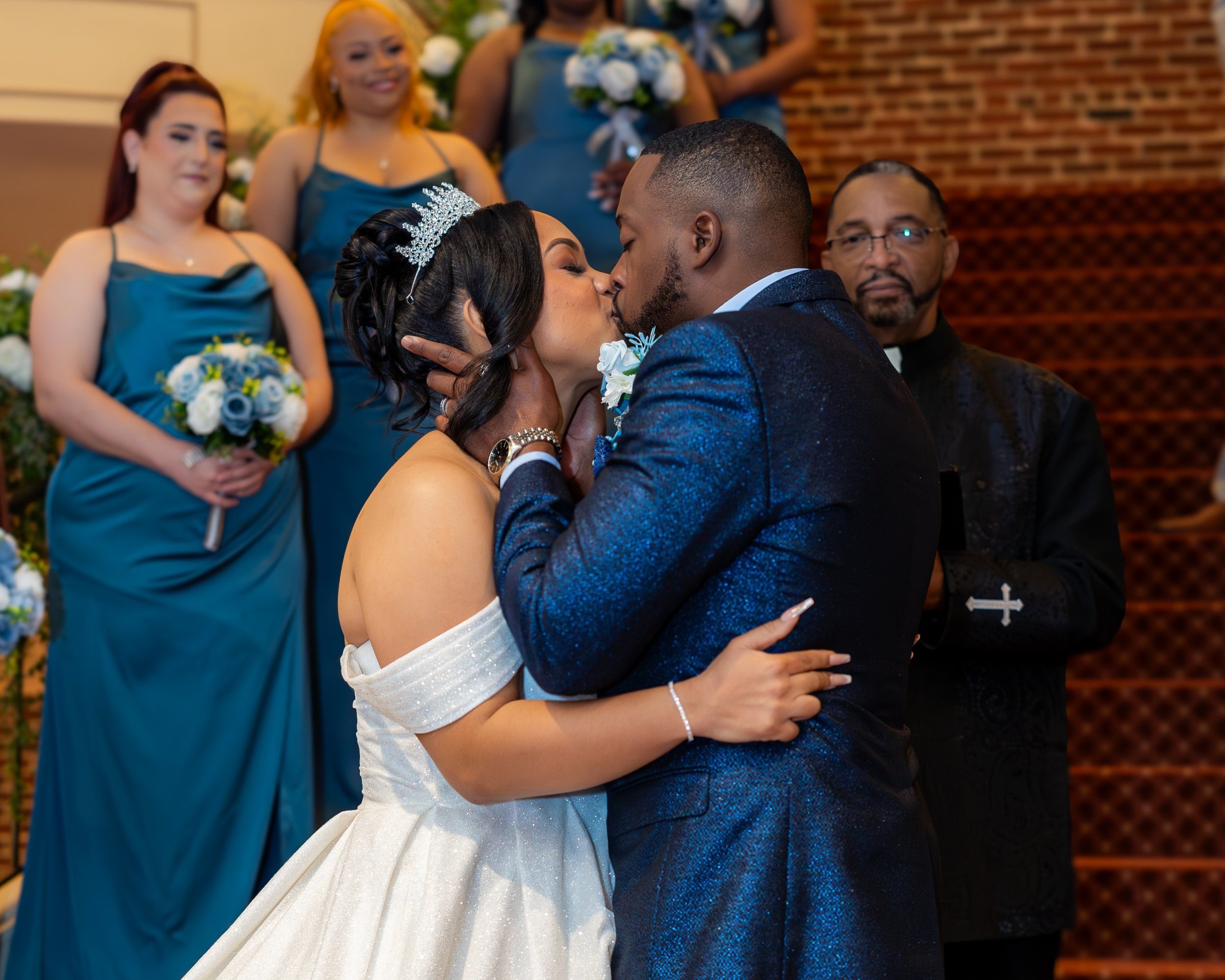 A newlywed couple shares a kiss during their wedding ceremony, surrounded by bridesmaids in blue dresses holding bouquets, and a officiant dressed in black, against a brick wall background.
