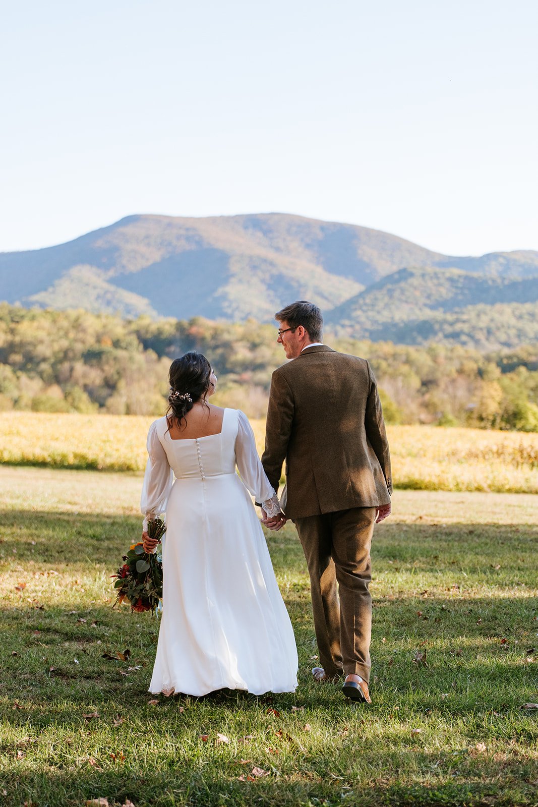 A bride and groom walking hand-in-hand in a field with mountains in the background. The bride is wearing a white wedding dress and holding a bouquet, and the groom is wearing a brown suit.