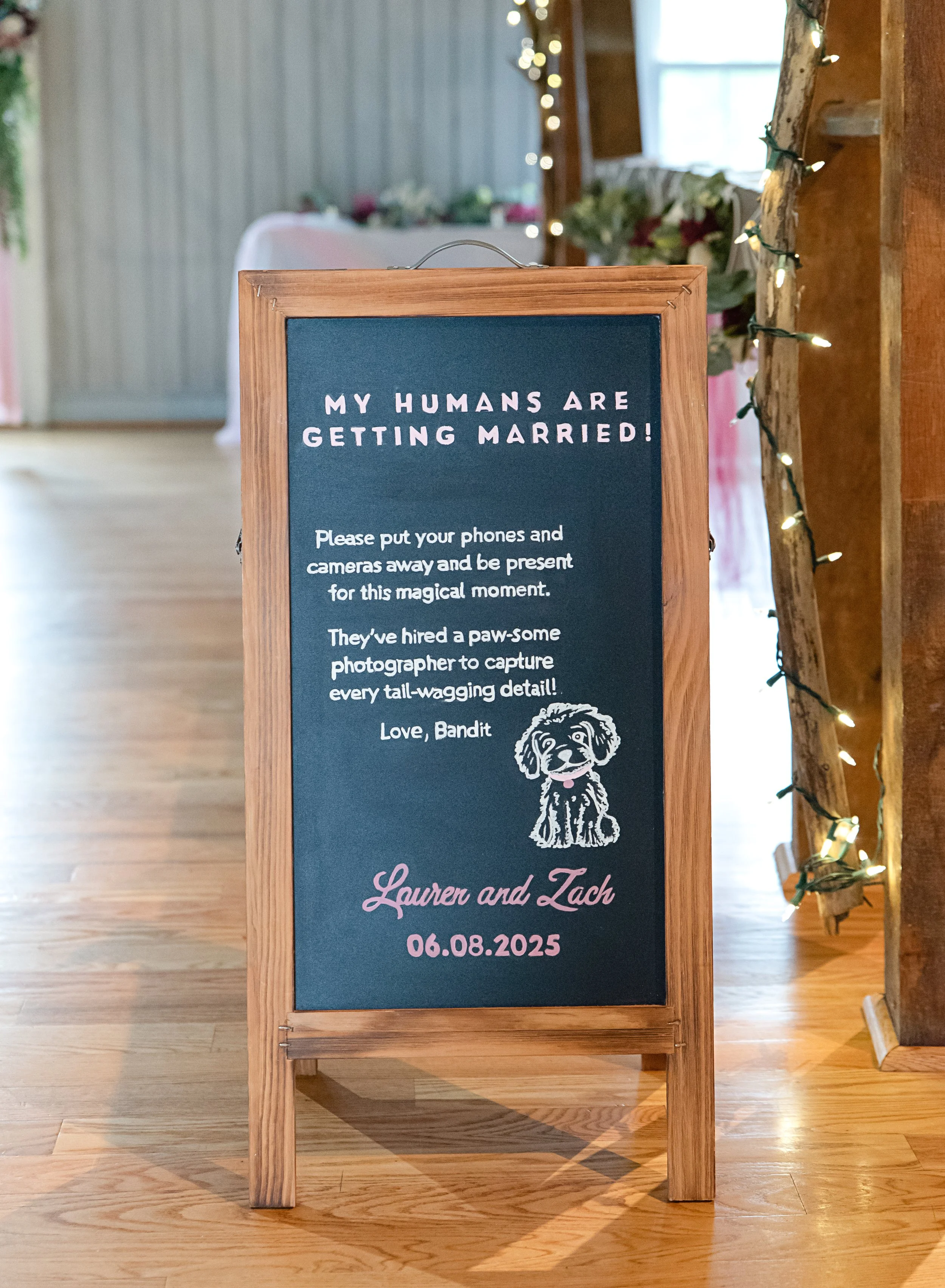 Wedding sign with a dark blue background and wooden frame, featuring a cute cartoon puppy and text about humans getting married on June 8, 2025, at a rustic venue decorated with fairy lights and flowers.