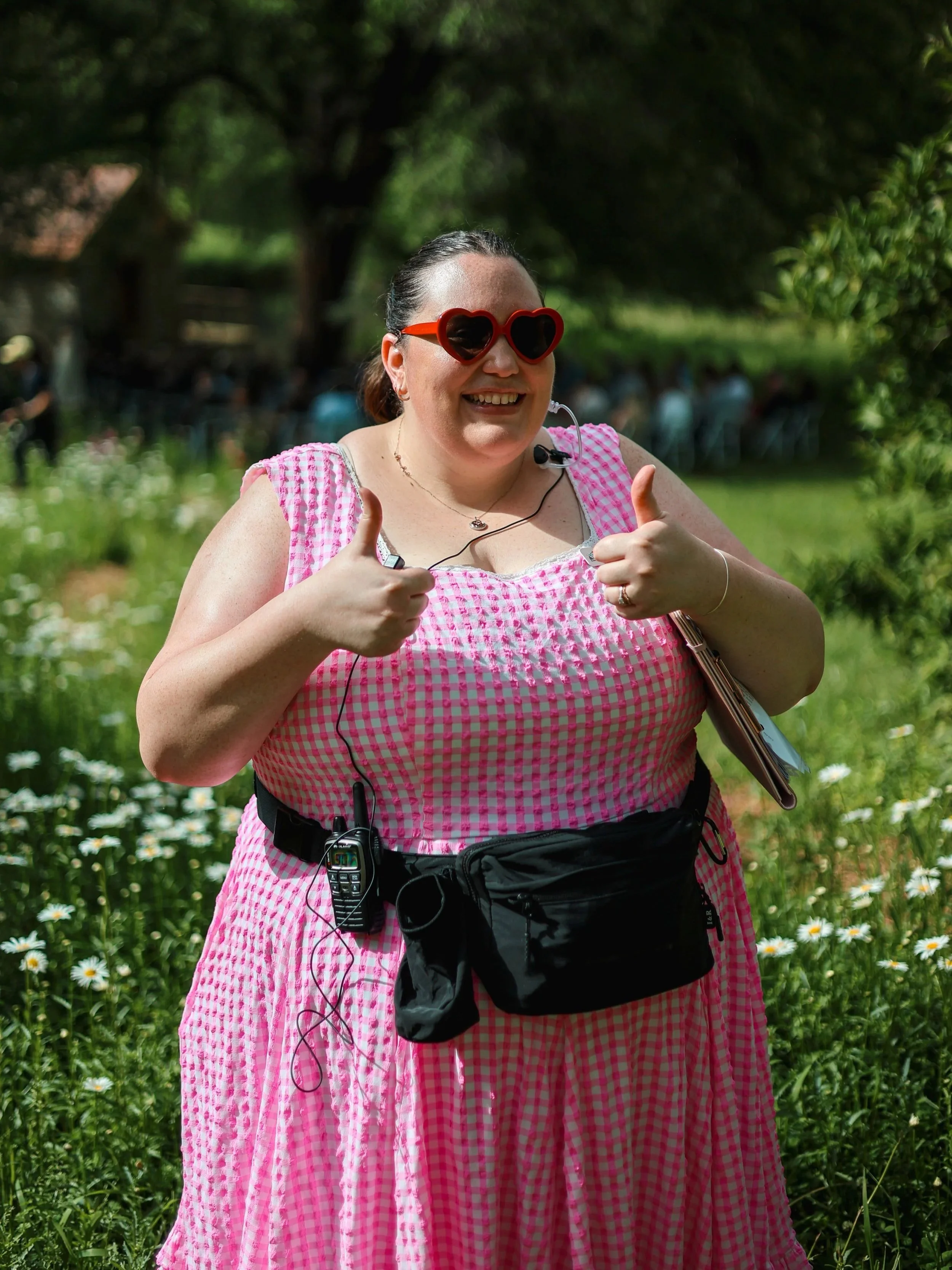 A woman wearing heart-shaped sunglasses and a pink checkered dress giving a thumbs up in a garden filled with daisies.