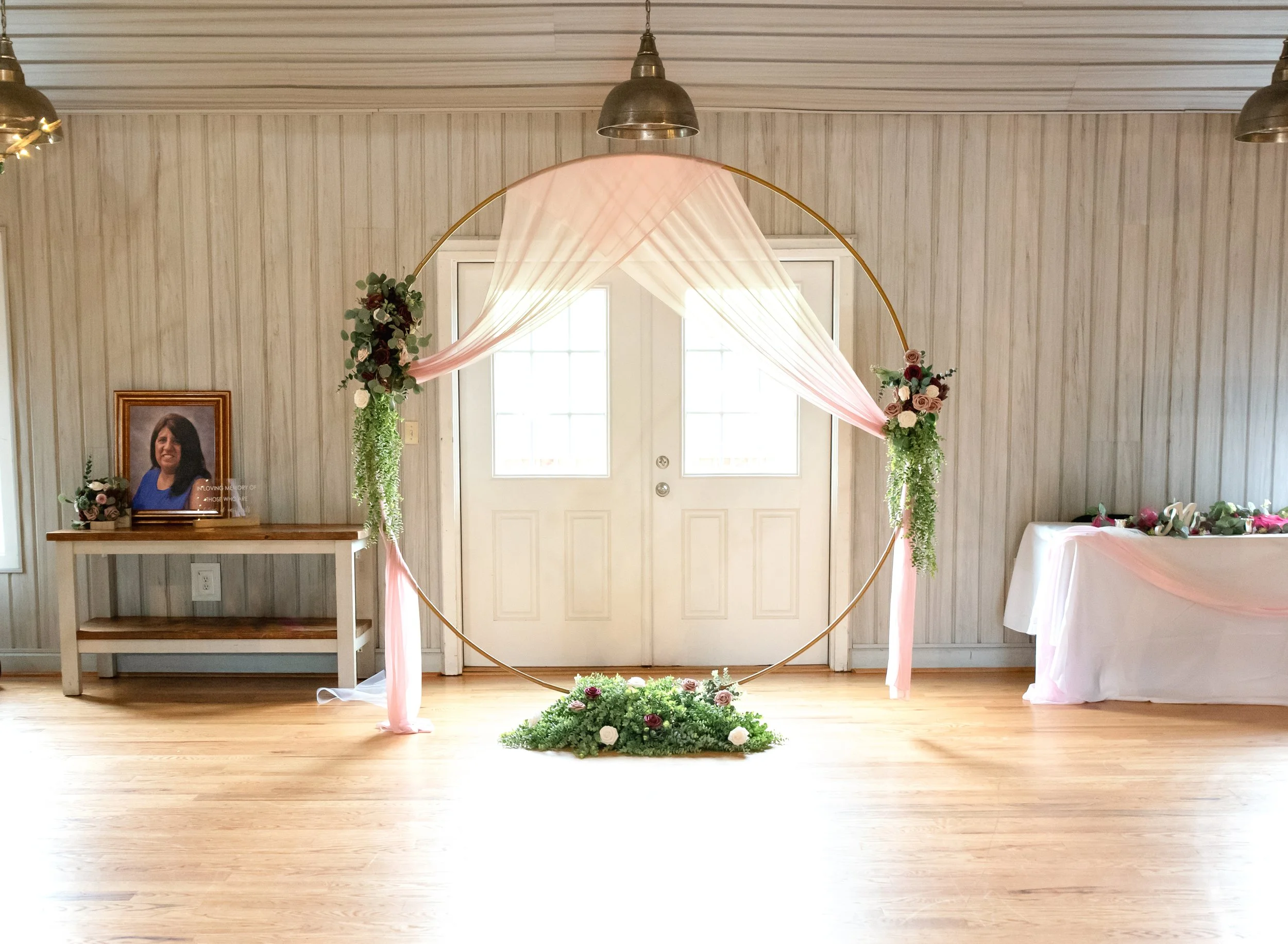 A decorated wedding aisle with a gold hoop arch draped in pink fabric and floral arrangements, set in a bright room with wooden floors and white walls.