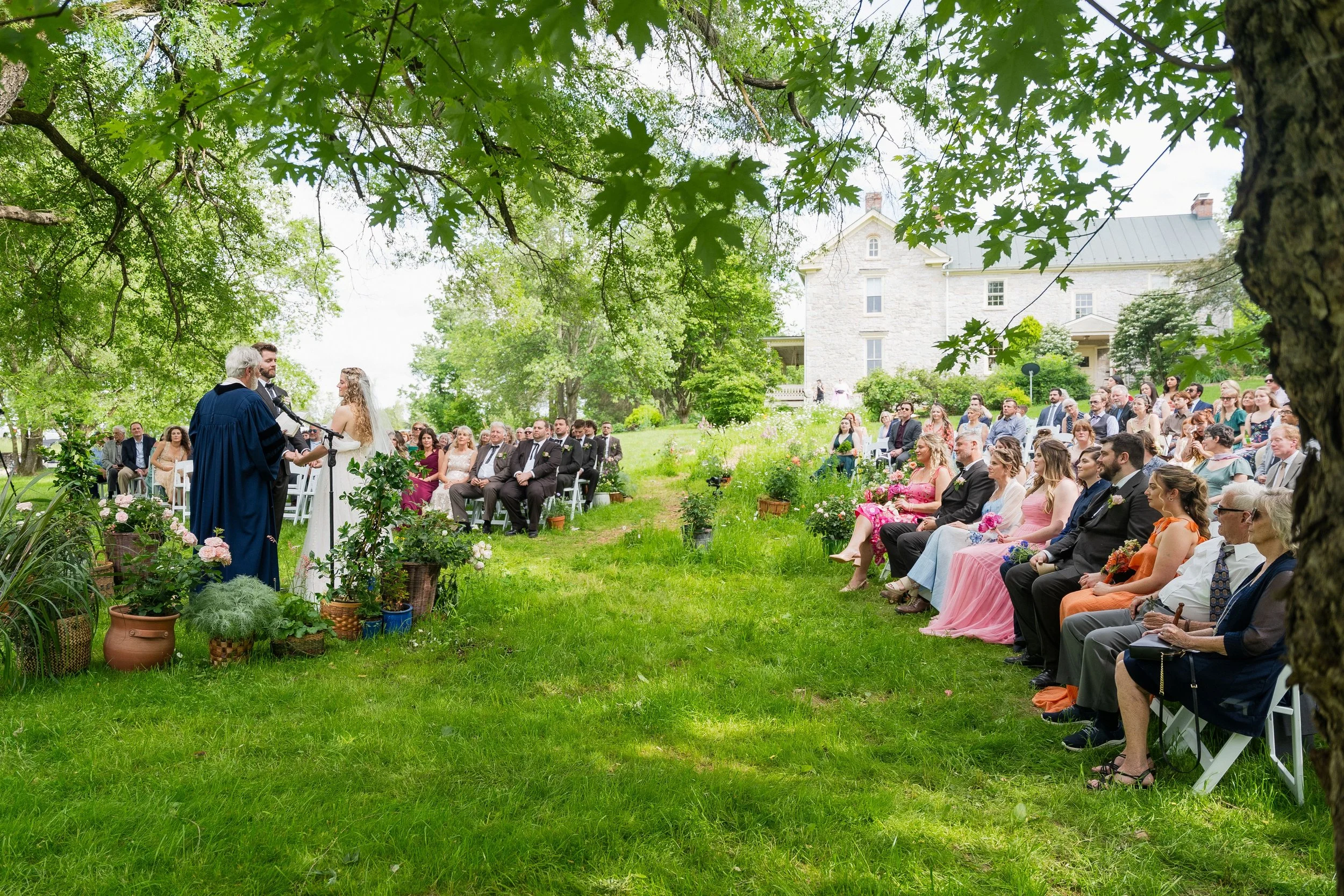 Wedding ceremony outdoors with bride and groom at the altar, officiant, and seated guests under green trees, in front of a large white house.