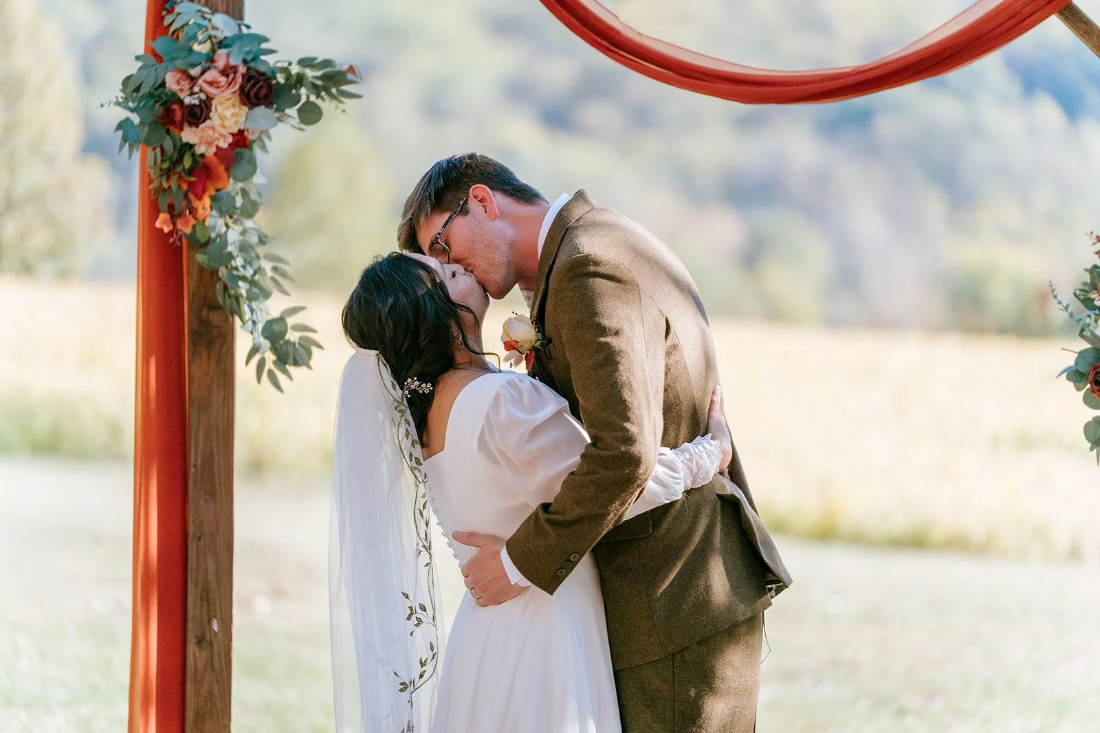 A couple kisses during an outdoor wedding ceremony. The woman wears a white dress and veil, and the man wears a brown suit. They are under a decorated arch with flowers and greenery.