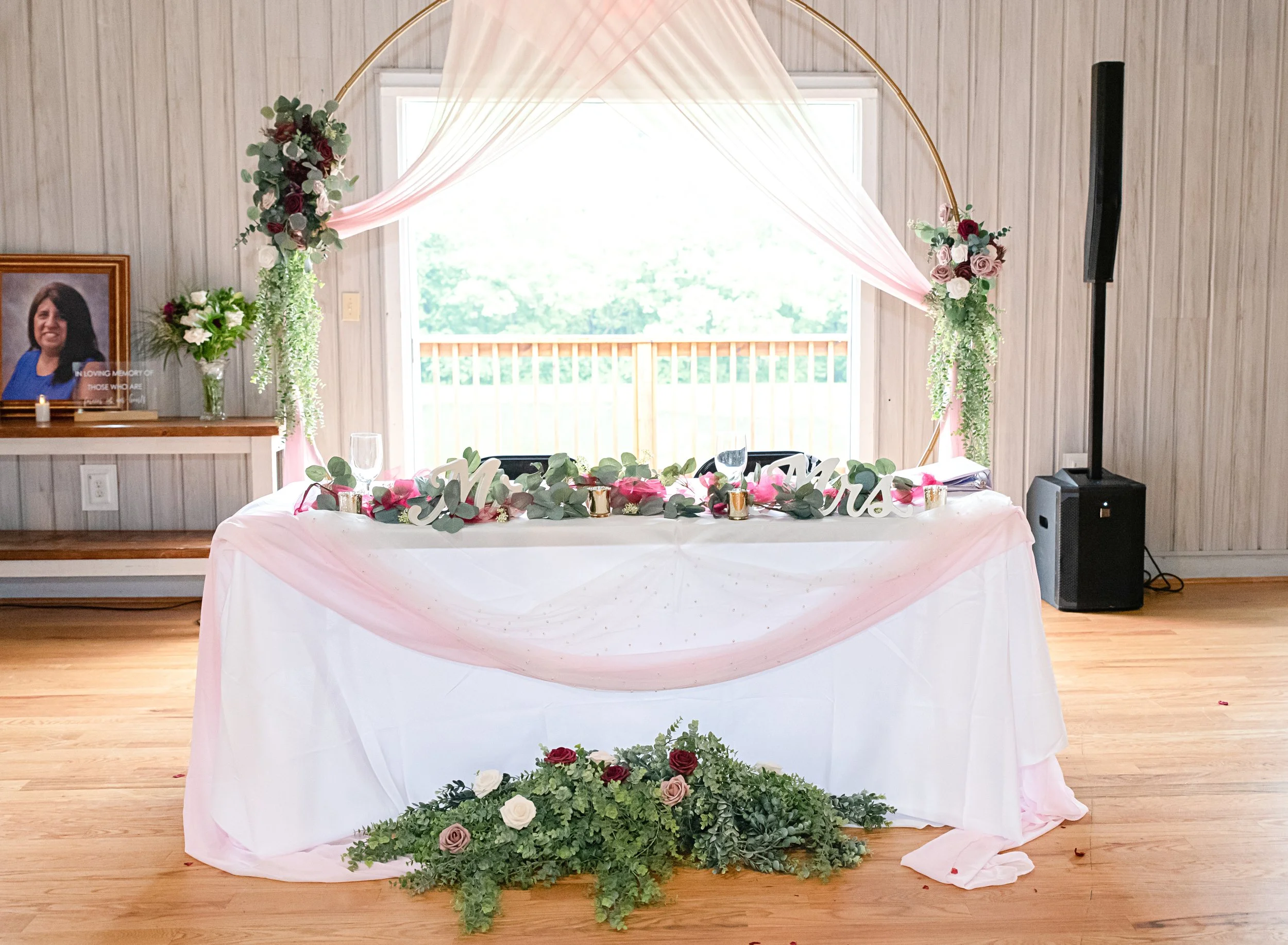 Decorated sweetheart table for a wedding reception with pink and green floral arrangements, a gold circle backdrop, and a pink sheer fabric draped over the table.
