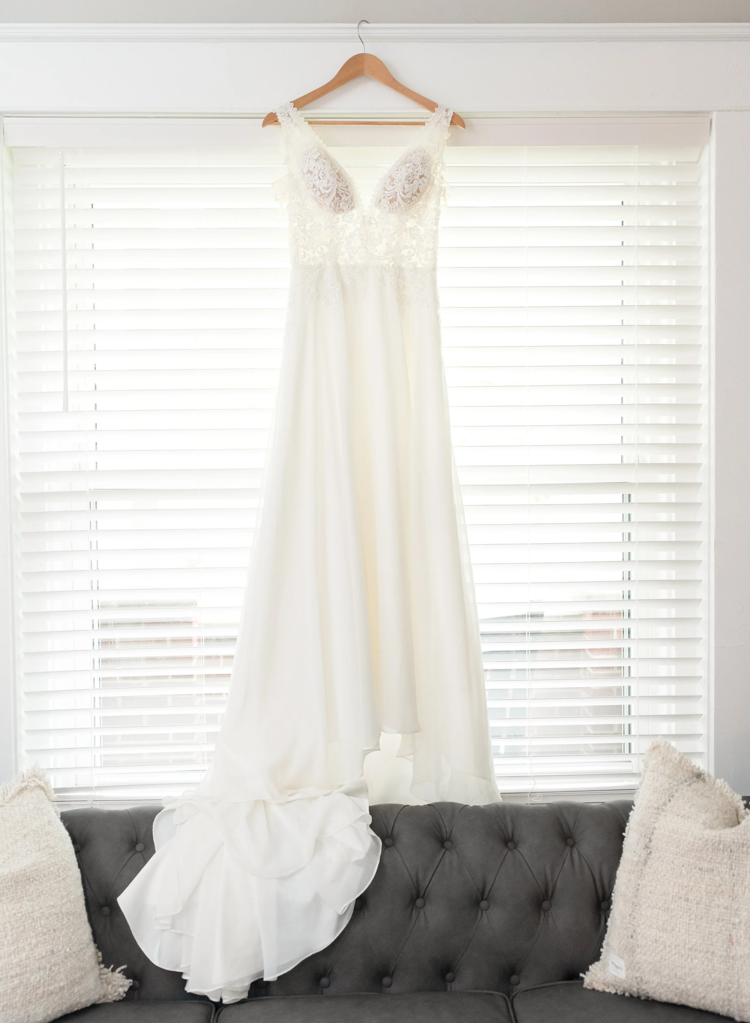Wedding dress hanging on a wooden hanger in front of a window with white blinds above a dark gray sofa with two white pillows.