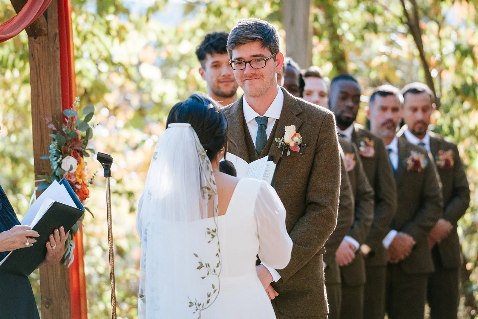 A wedding ceremony taking place outdoors, with a bride and groom exchanging vows as a woman officiates. Several groomsmen stand in the background, and the scene is decorated with flowers and greenery.