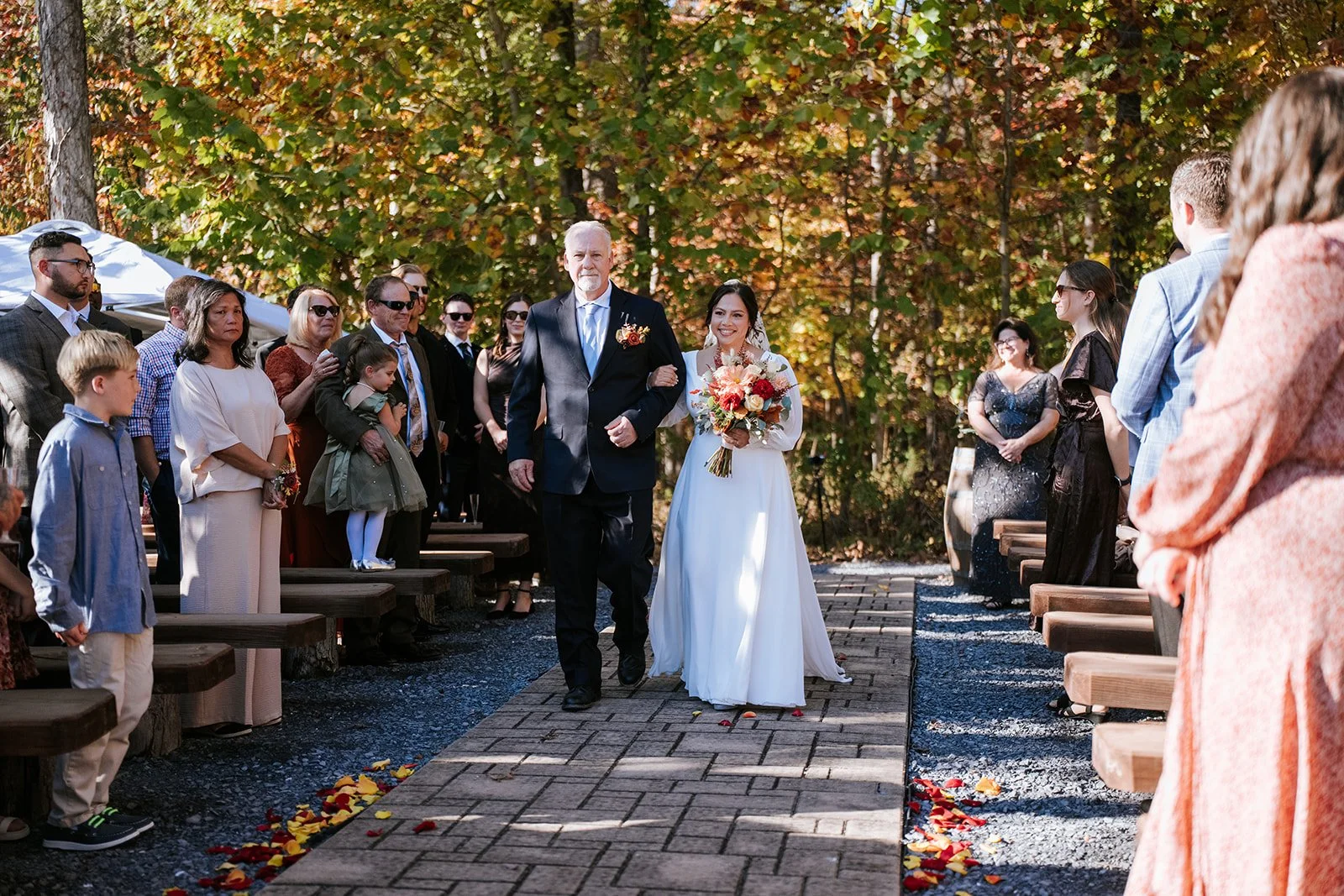 A bride walking down the aisle with an older man, likely her father, at an outdoor wedding ceremony surrounded by friends and family, with autumn trees in the background.