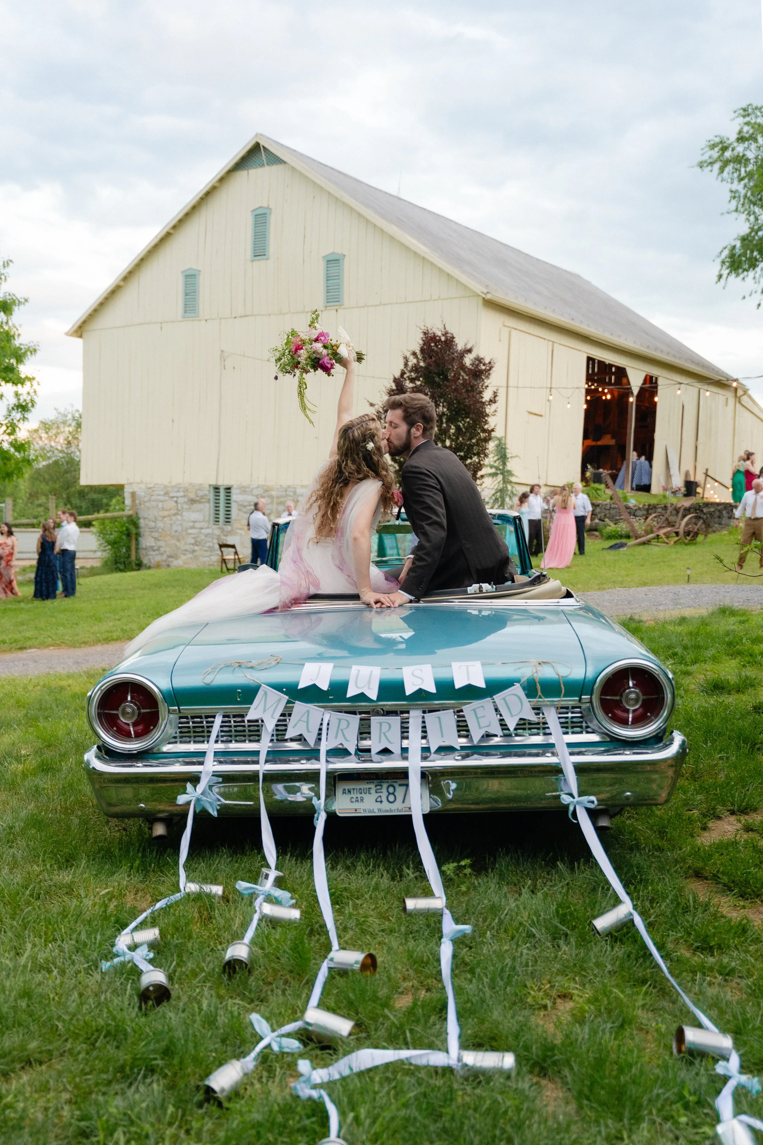 A newlywed couple kissing in a vintage blue convertible car with a 'Just Married' banner and ribbons, outdoors near a barn with wedding guests in the background.