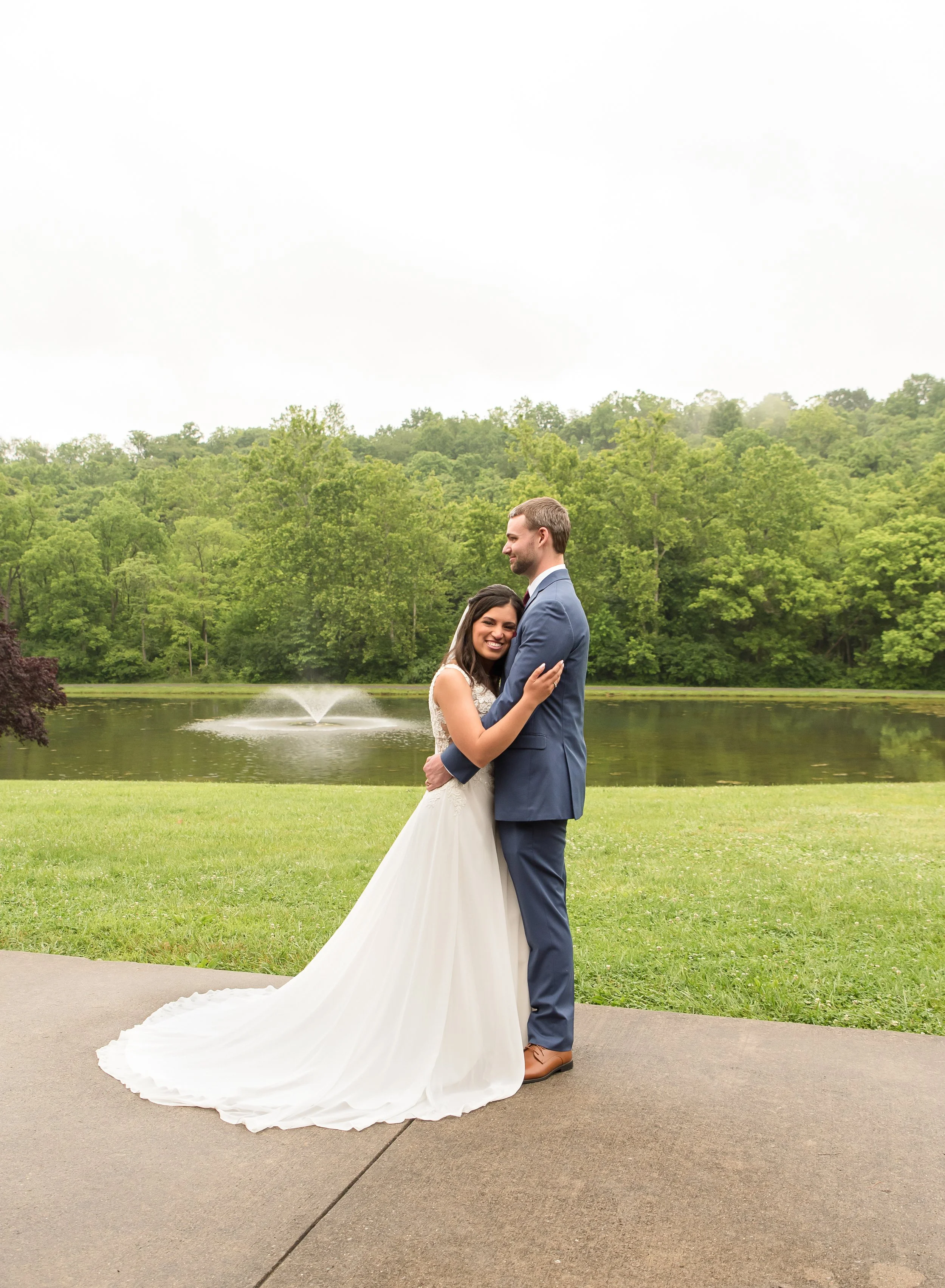 A couple in wedding attire standing happily near a pond with fountain and surrounded by green trees