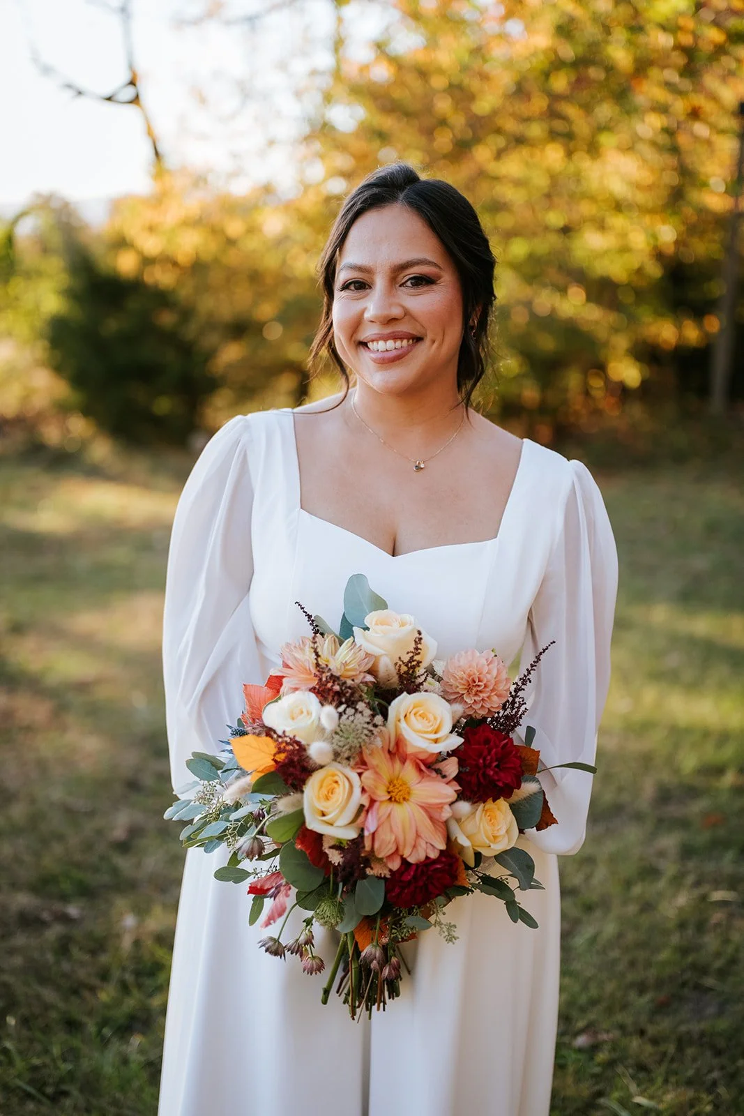 Happy bride in a white wedding dress holding a bouquet of white, peach, red, and pink flowers outdoors during autumn.