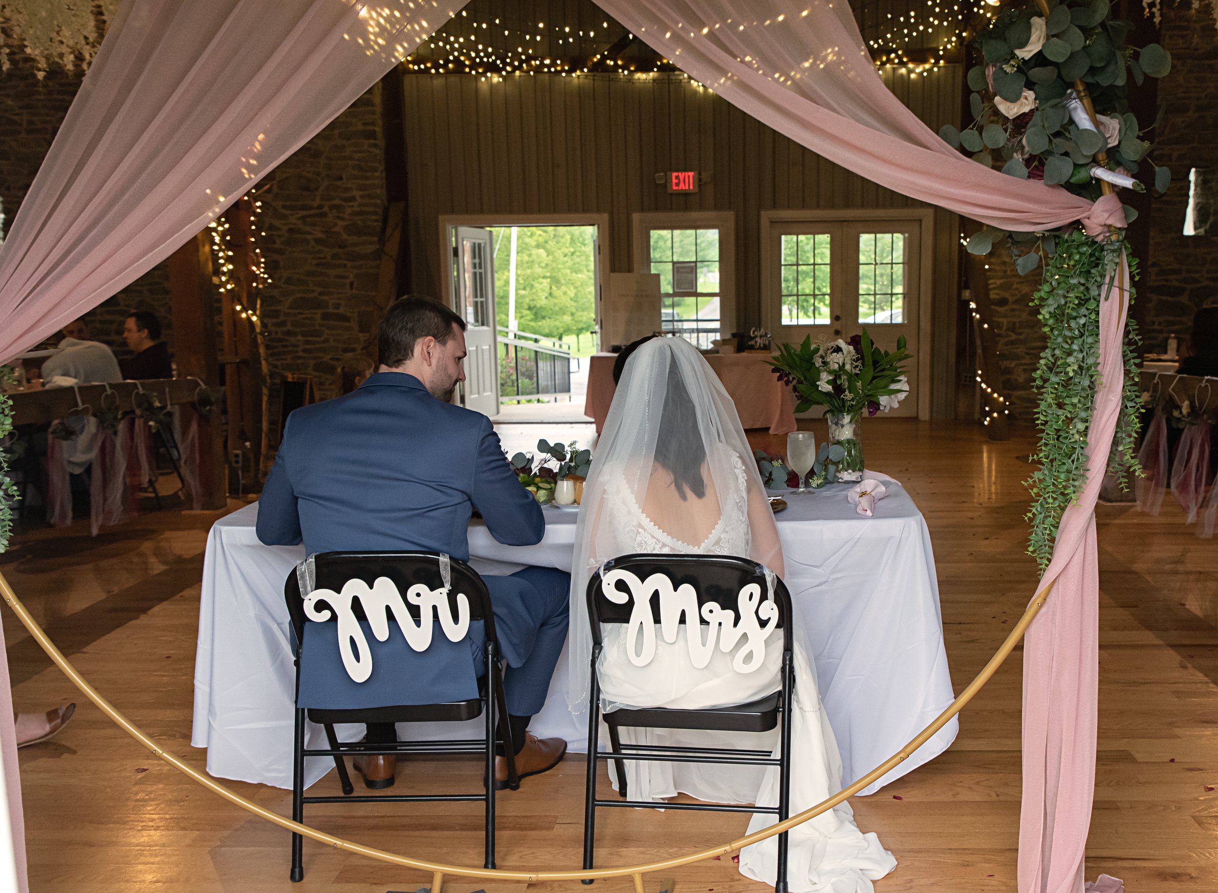 Newlyweds sitting at a decorated reception table under a pink draped canopy with fairy lights, with "Mr" and "Mrs" signs on the chairs, in a rustic venue.