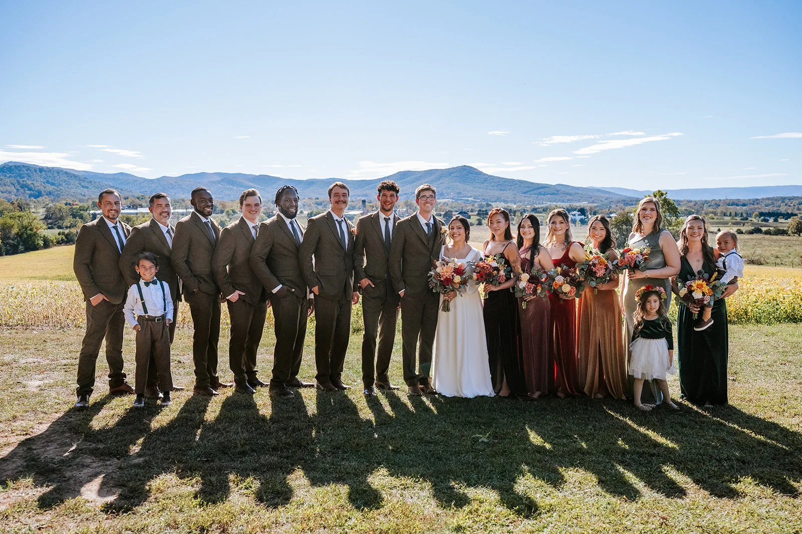 Wedding party standing outdoors in a field with mountains in the background. The group includes men in suits, women in dresses holding bouquets, children, and a bride in a white dress. They are smiling and posing for a photo on a sunny day.