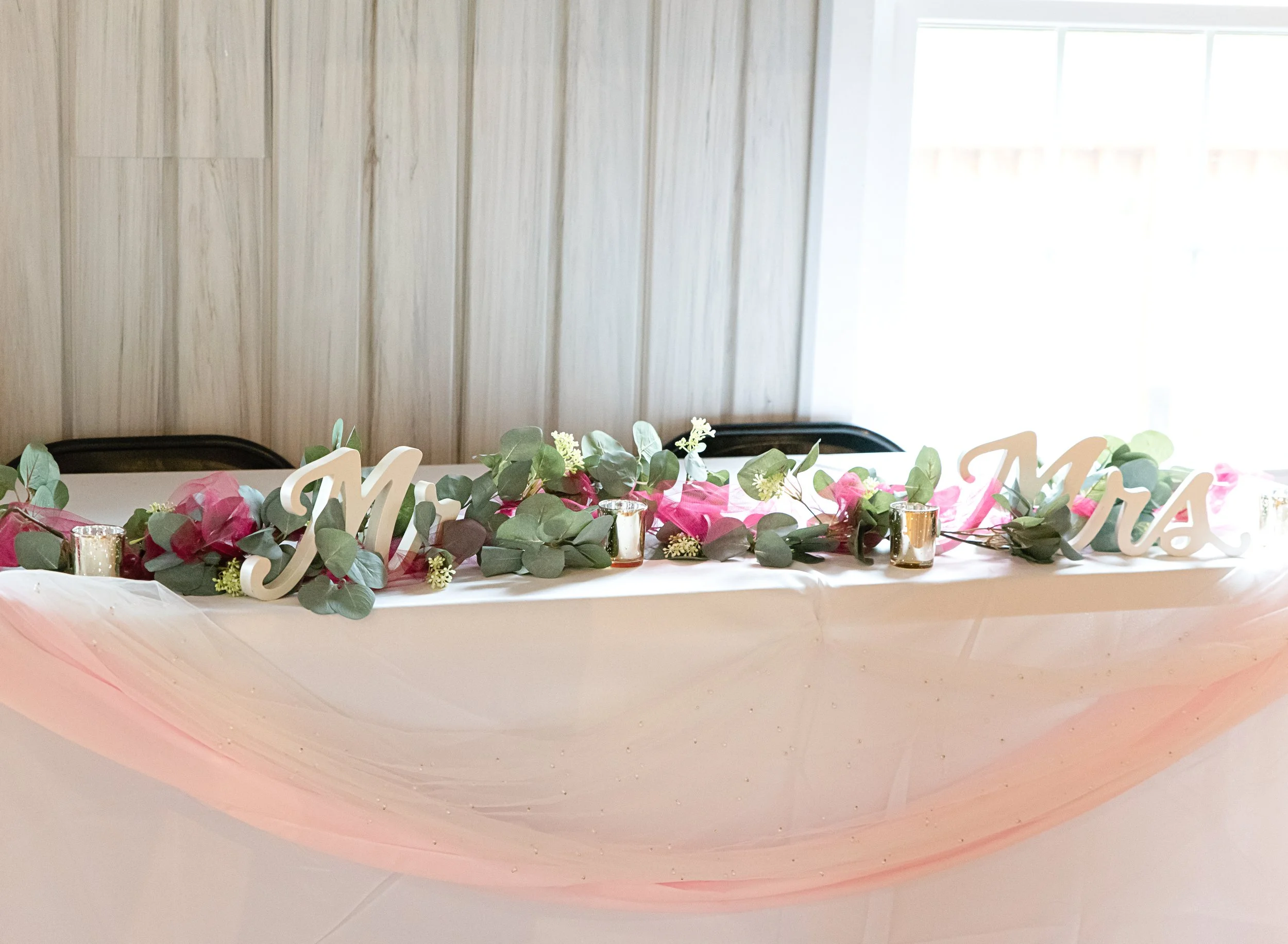 Wedding table decorated with pink flowers, green foliage, 'Mr & Mrs' signs, and small candles, with a white tablecloth and pink tulle decoration.