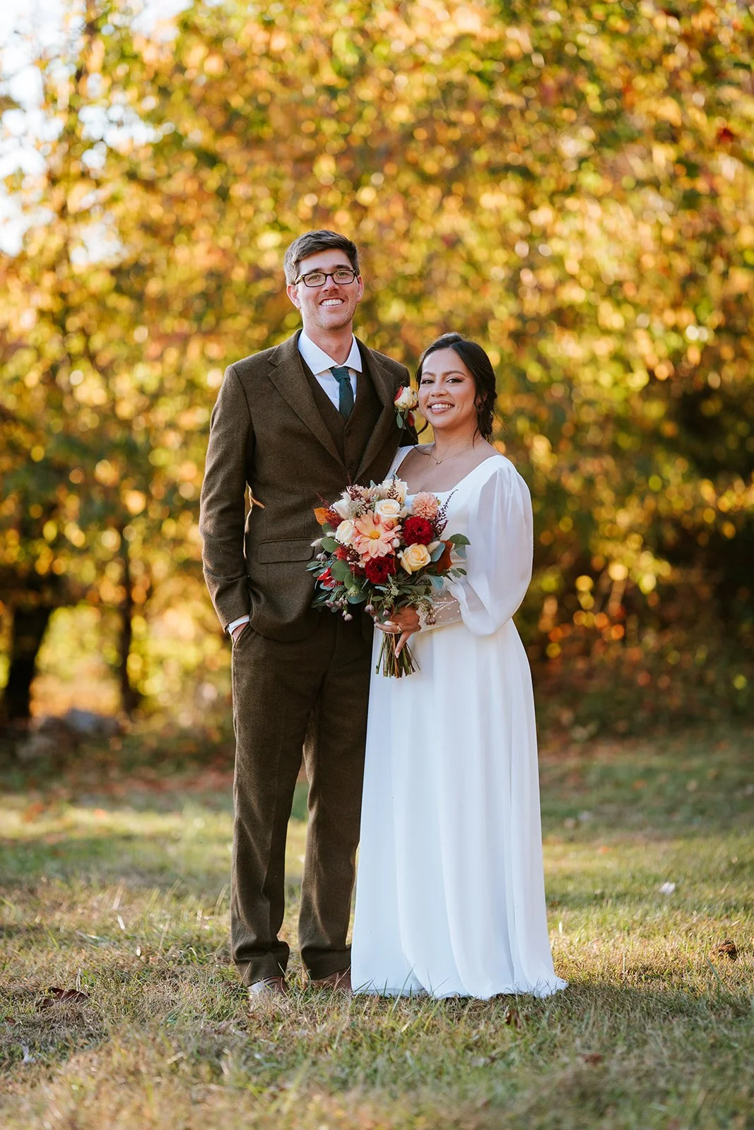 A couple standing outdoors in autumn, dressed in wedding attire, with the woman holding a bouquet of flowers