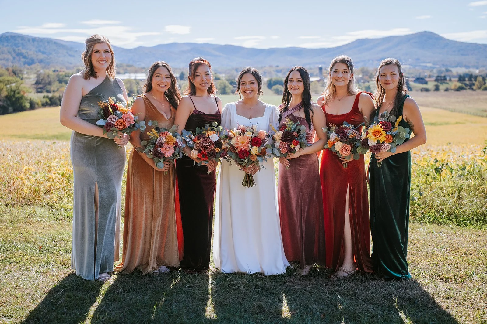 A group of seven women standing outdoors in a field with mountains in the background, all holding bouquets of flowers, dressed in colorful and elegant dresses for a special occasion.