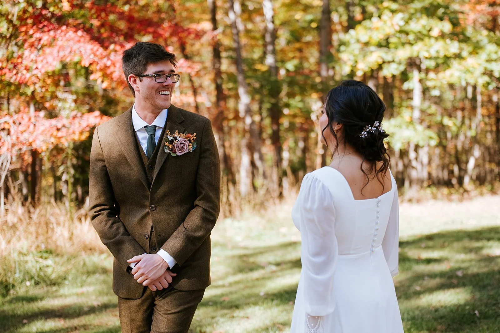 A couple getting married outdoors in a park with fall foliage in the background. The groom is wearing a brown suit with a floral boutonniere, and the bride is in a white dress with buttons on the back, facing each other and smiling.