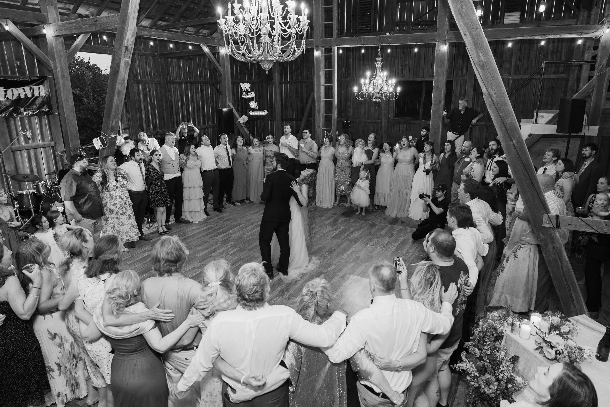 A wedding reception in a rustic barn with a couple dancing in the center surrounded by guests. The barn features wooden walls, chandeliers, and string lights.