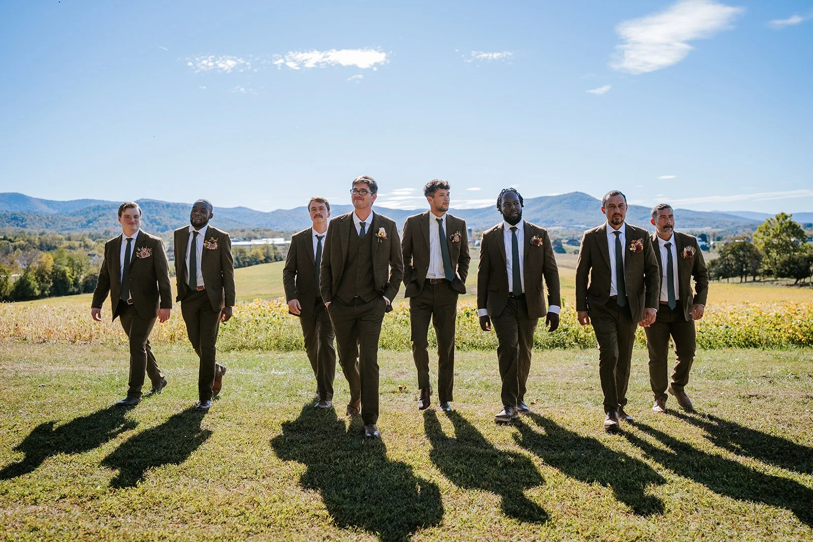 A group of nine men dressed in brown suits and ties, walking outdoors on a grassy field with mountains and trees in the background under a blue sky.