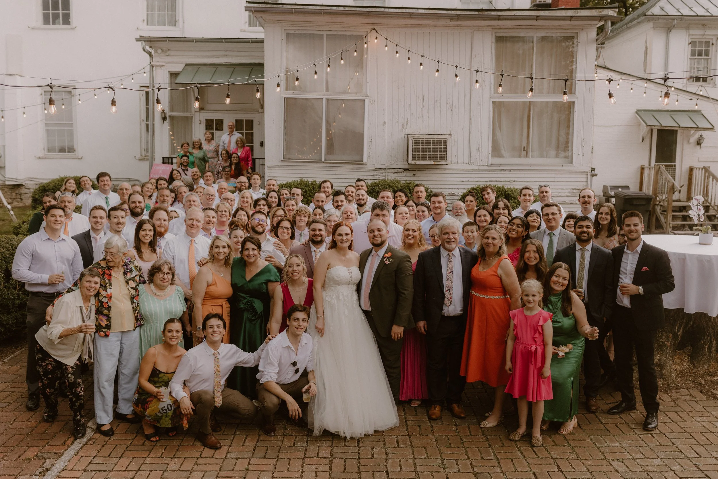 A large group of people gathered outside a white building with string lights hanging overhead for a wedding celebration. The bride and groom are in the front center, smiling, with the bride in a white wedding dress and the groom in a suit. The group 