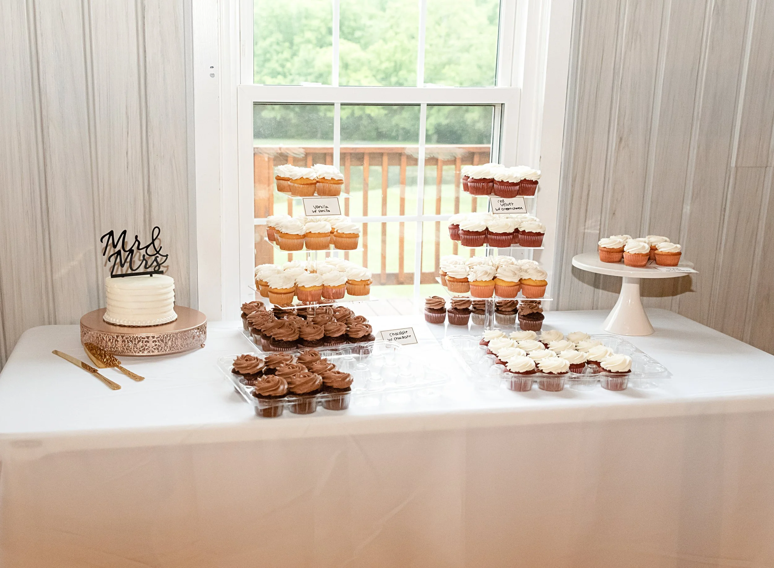 A dessert table featuring a small white frosted wedding cake with 'Mr & Mrs' topper, decorated with cupcakes in chocolate, vanilla, and red velvet flavors, displayed on multi-tiered stands and a tray, with a window in the background.