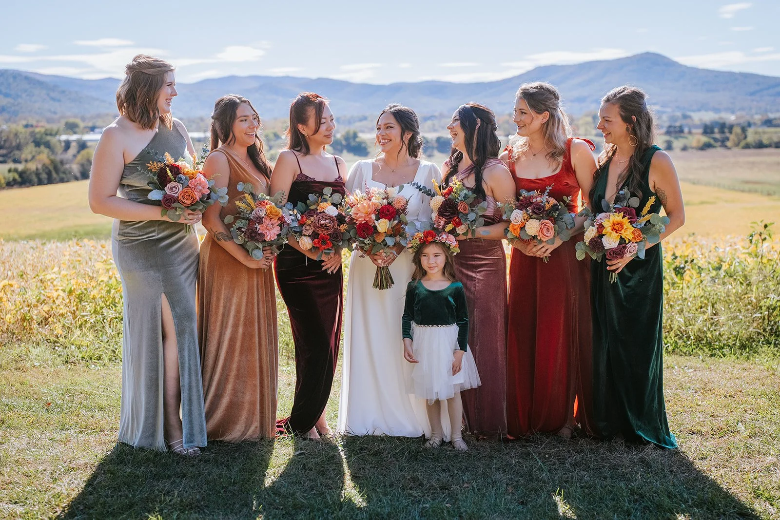 Women and a girl at a wedding outdoors, holding bouquets of flowers, standing in a field with mountains in the background.