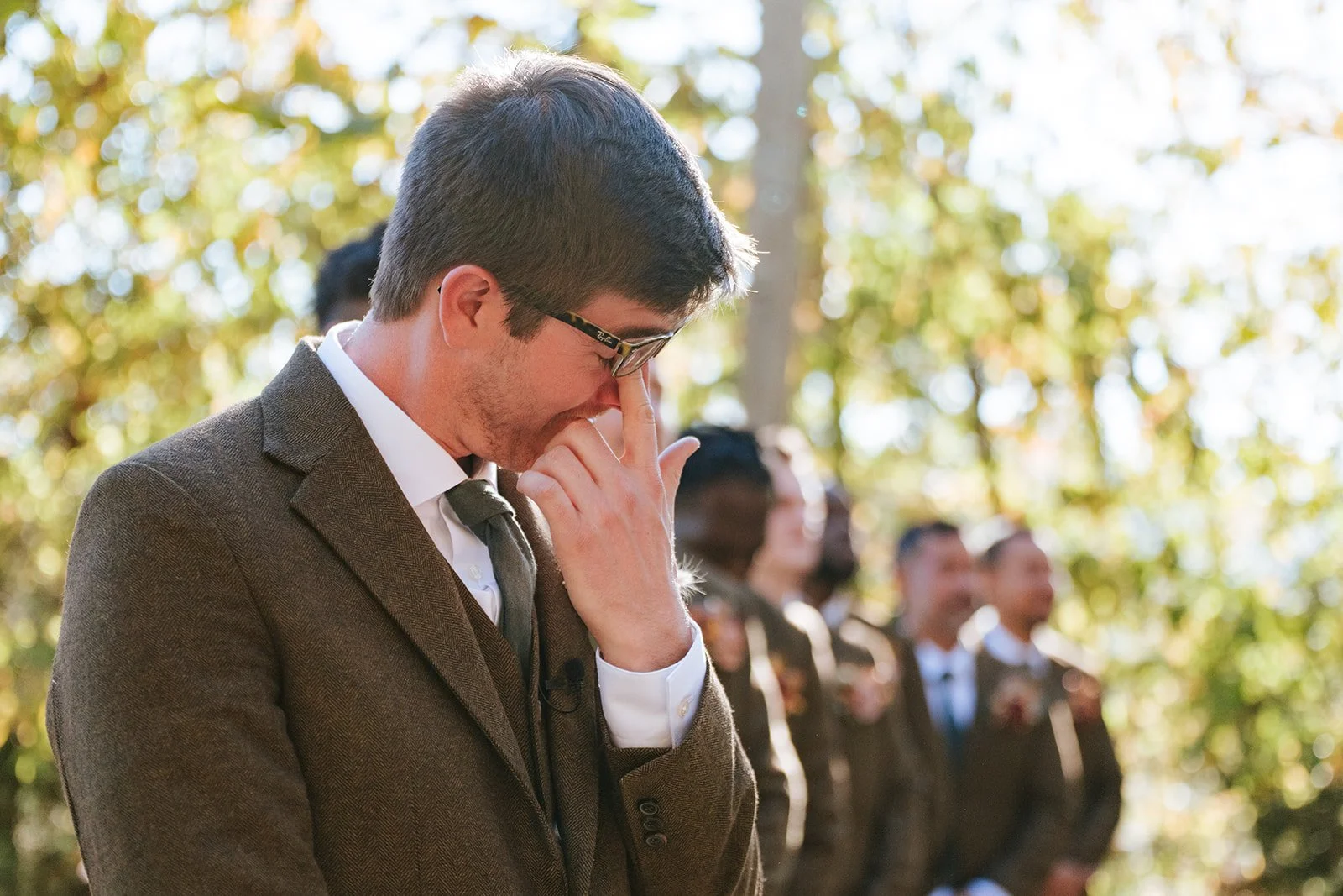 A man in a brown suit and glasses is holding his face with his hand, with his eyes closed, during an outdoor ceremony with other people dressed formally behind him.