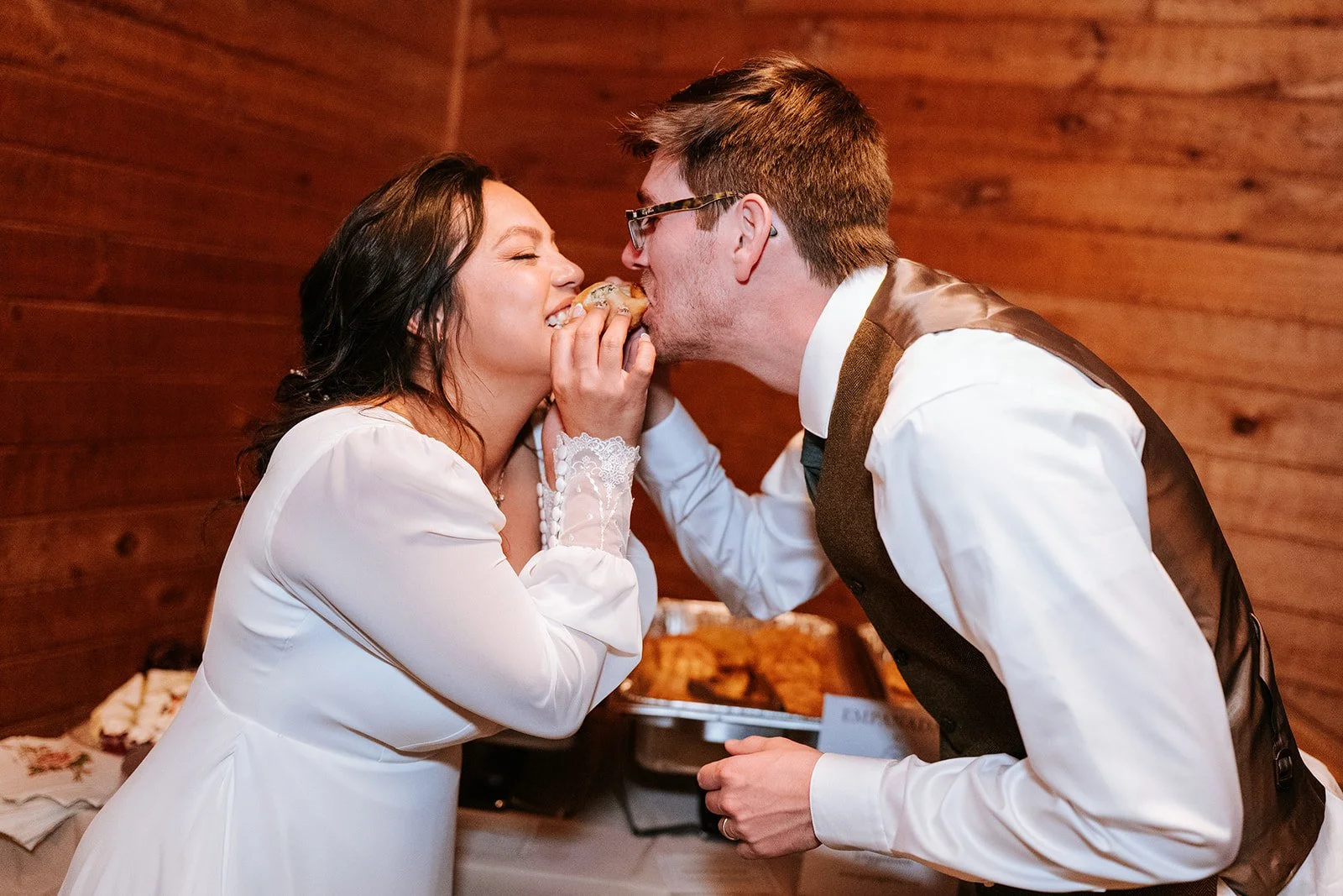 A bride and groom share a playful moment as the groom feeds the bride a piece of food during their wedding reception, with a wooden wall in the background.
