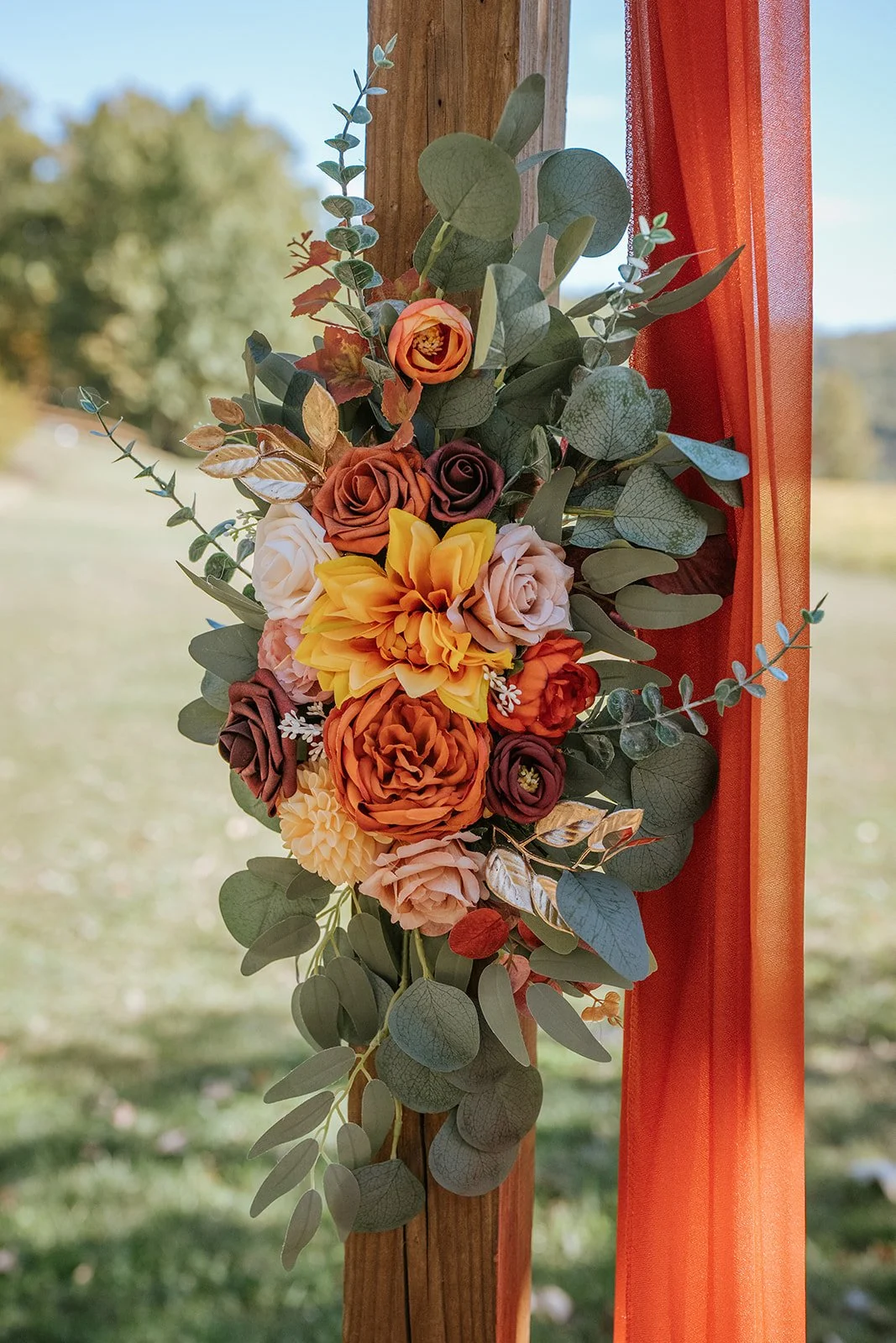 Bouquet of artificial flowers including roses, dahlias, and greenery attached to a wooden post with orange fabric, outdoors with blurred trees in the background.