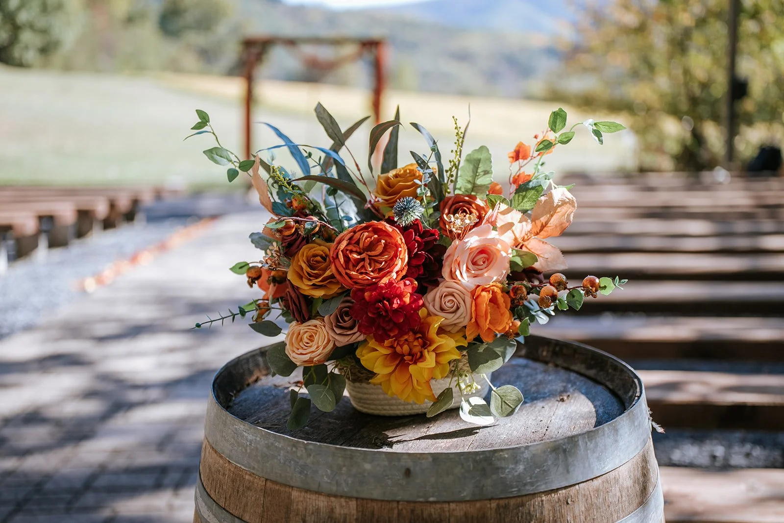 A vibrant bouquet of orange, peach, red, and yellow roses and flowers with green foliage in a wicker basket, placed on top of a wooden barrel outdoors with a blurred background of a picnic shelter and trees.