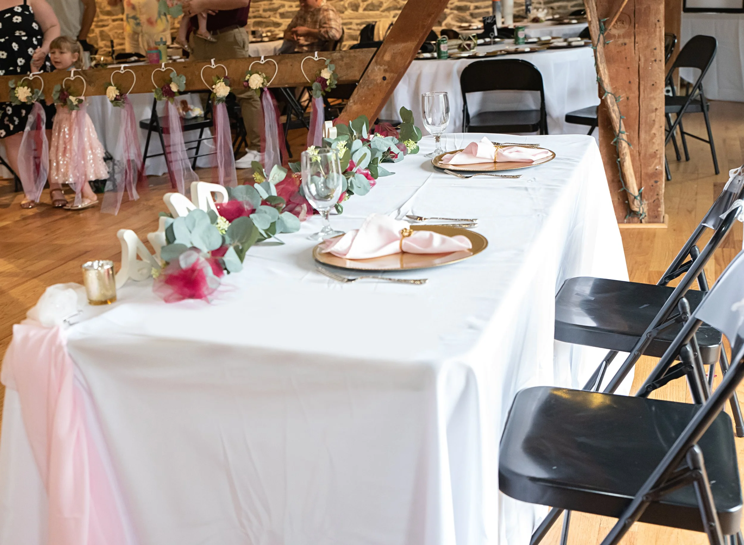Wedding reception table with white tablecloth, pink napkins on gold plates, glassware, and a floral centerpiece with eucalyptus leaves and pink flowers. In the background, children and adults near a decorated wooden beam with hanging flower hearts an