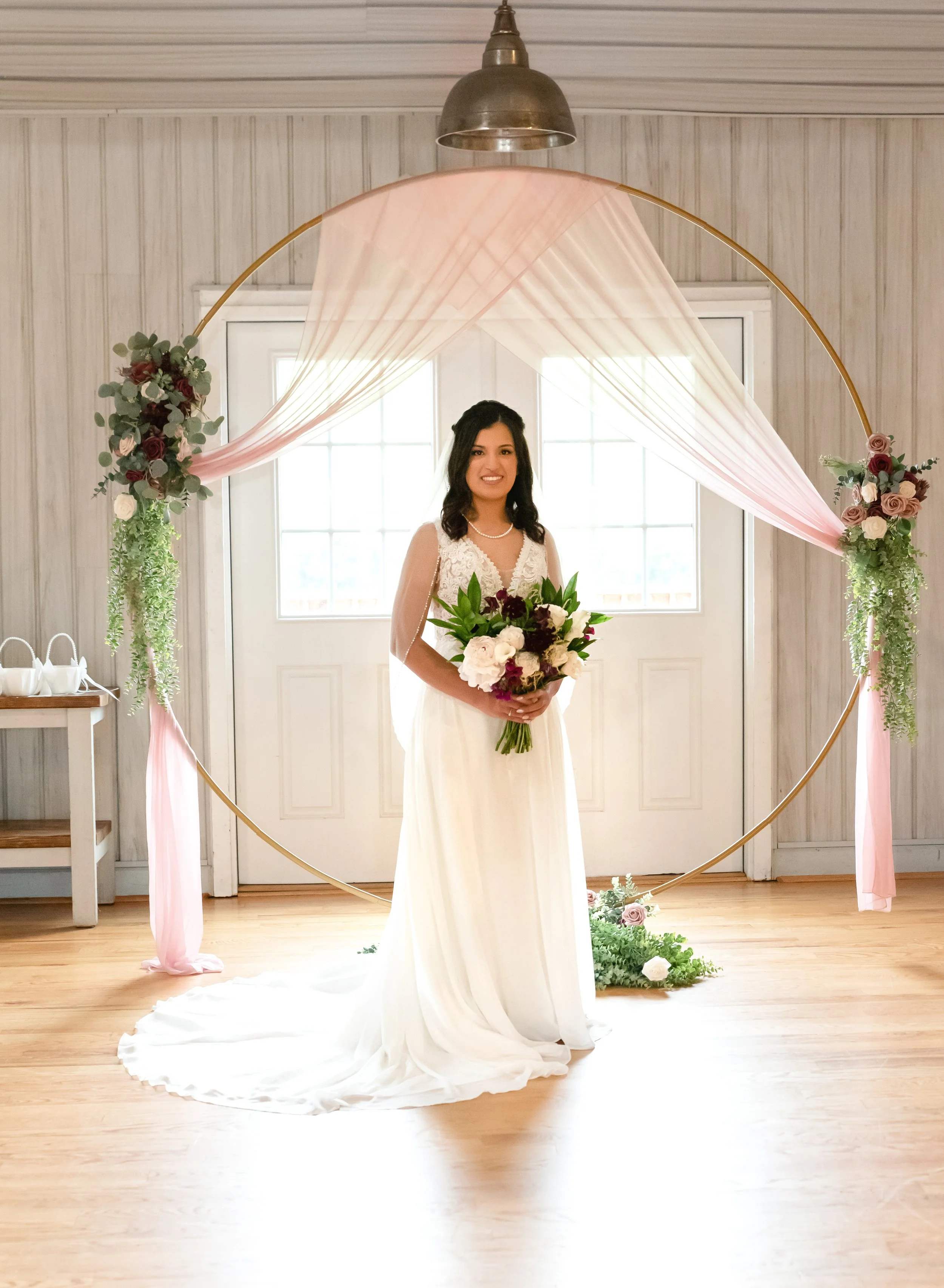 A woman in a white wedding dress holding a bouquet of flowers standing in front of a circular floral arch decorated with pink and purple roses and greenery, inside a bright room with white walls and wooden floor.