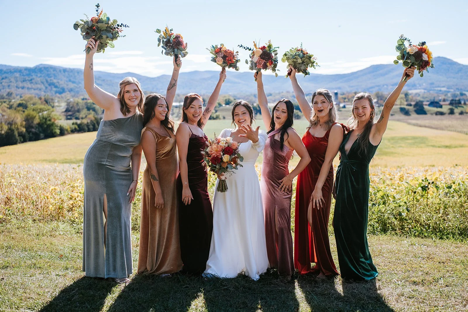 A group of women in colorful dresses celebrating outdoors on a sunny day, holding bouquets and raising them in the air, with mountains and a field in the background.