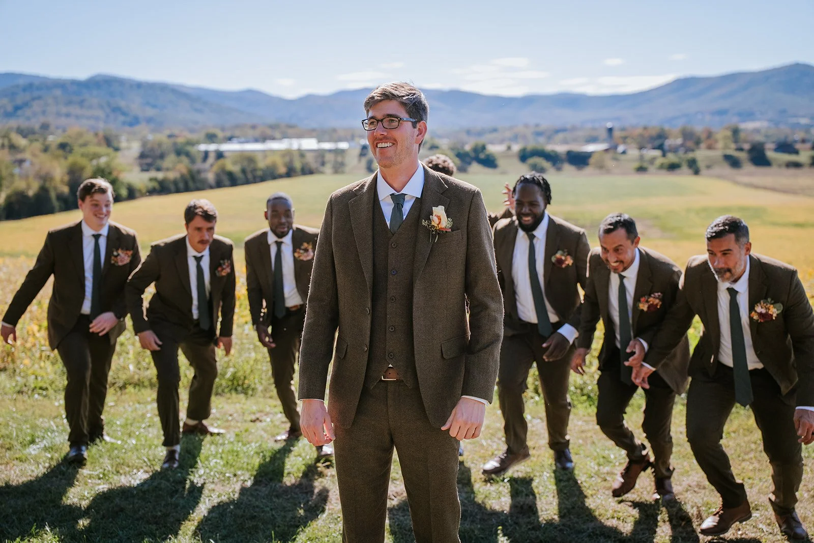A groom in a brown suit smiling in front of his wedding party, with seven groomsmen in suits standing outdoors on a sunny day in a grassy field with mountains in the background.