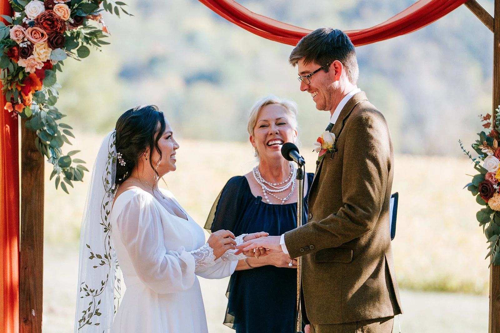 A bride and groom holding hands during their outdoor wedding ceremony, exchanging vows, with a officiant standing between them and smiling, framed by a floral arch with roses and greenery.