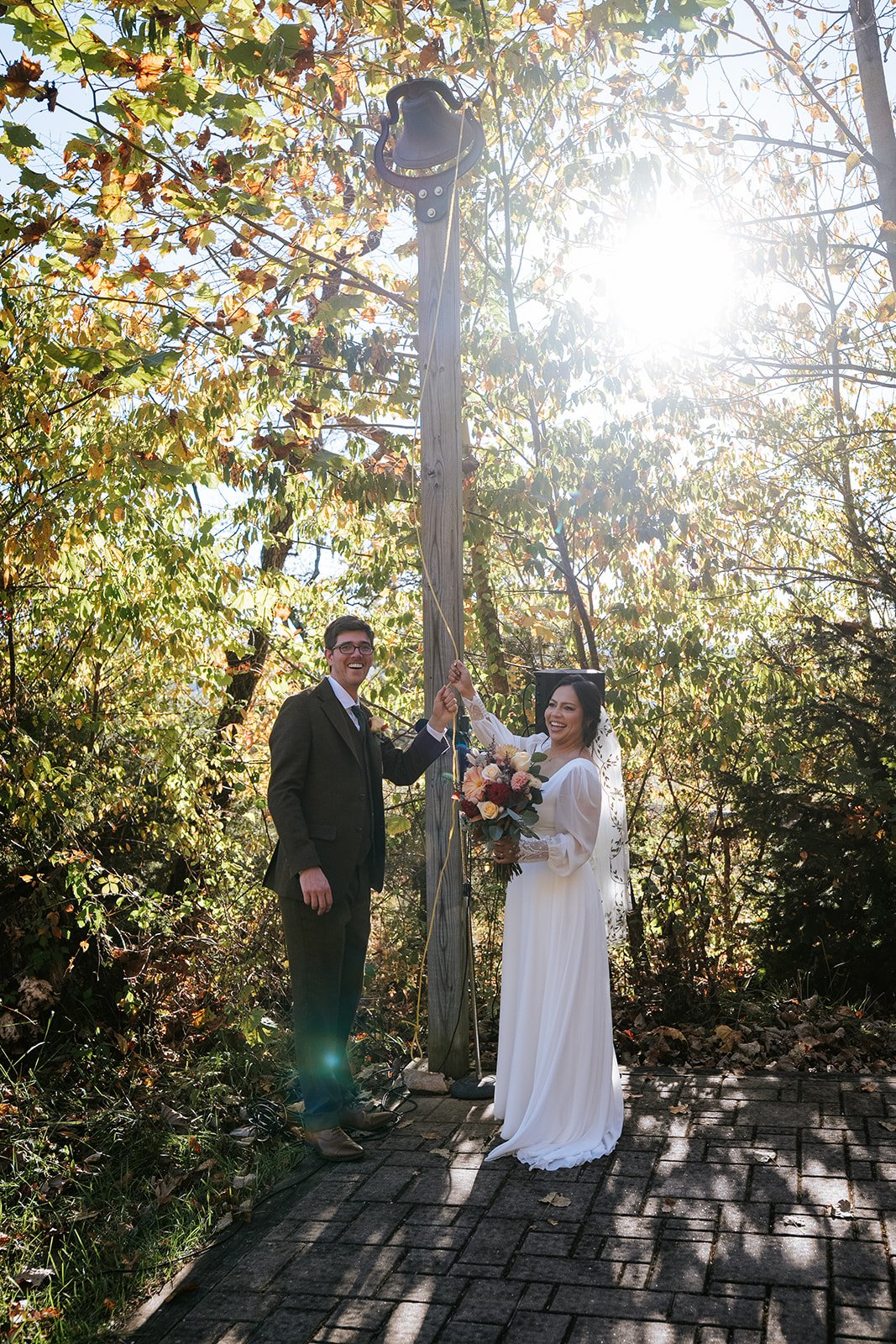 A newlywed couple in wedding attire smiling and holding hands under a utility pole with a bell and speaker, among autumn-colored trees with sunlight filtering through.
