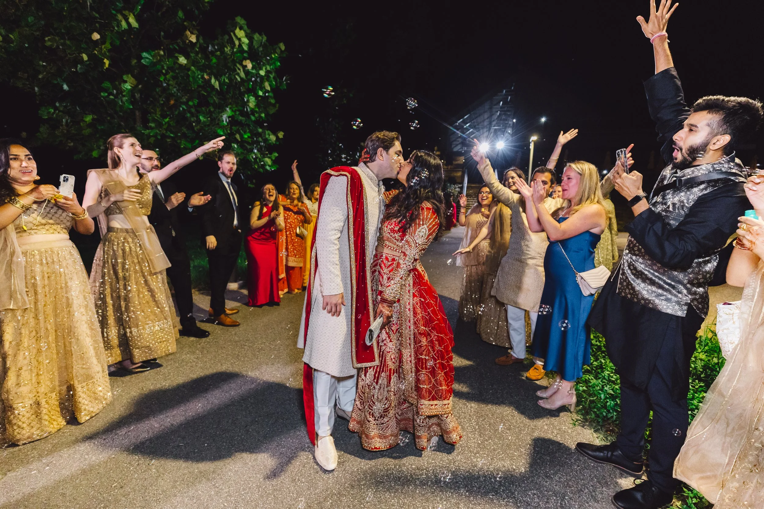 A couple dressed in traditional Indian wedding attire sharing a kiss surrounded by guests celebrating at night.