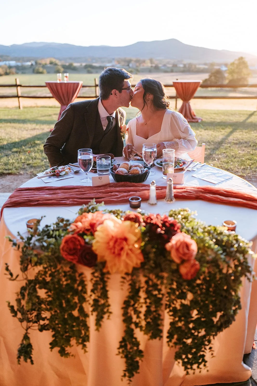 A couple is kissin' at an outdoor wedding reception, with mountains and a sunset in the background, and a table with flowers, food, and drinks in the foreground.