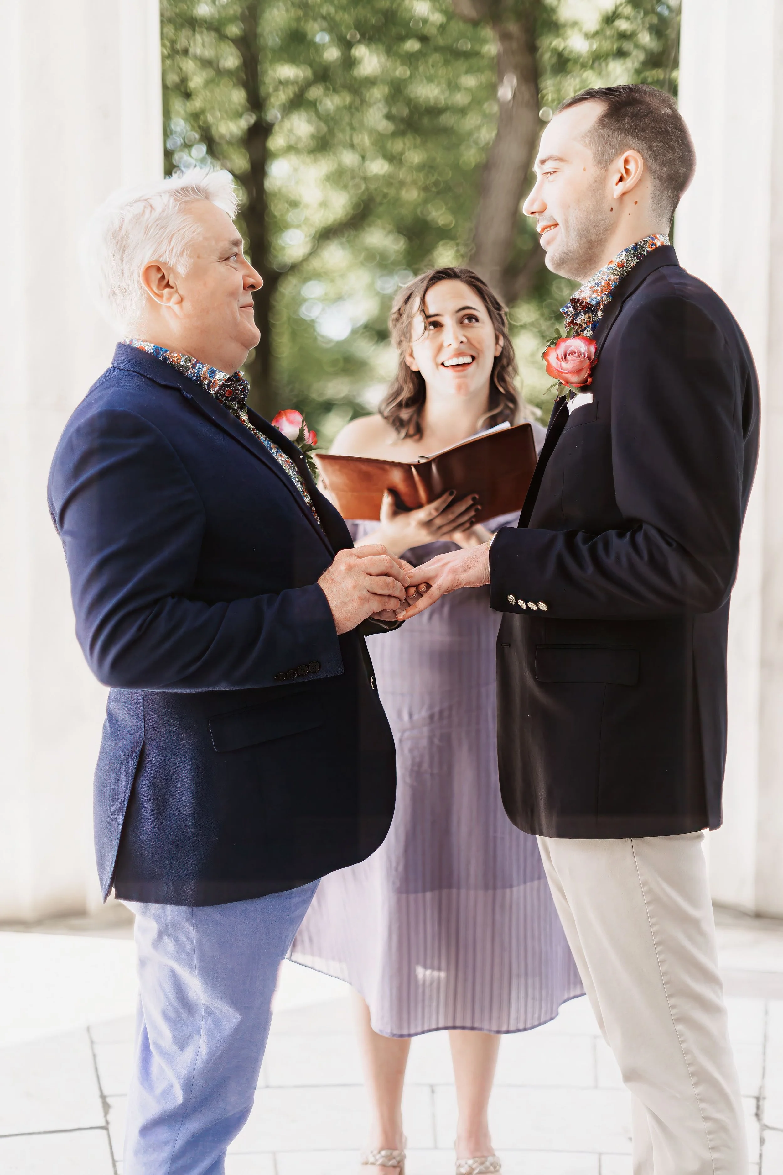 Two men exchanging rings during an outdoor wedding ceremony with an officiant.